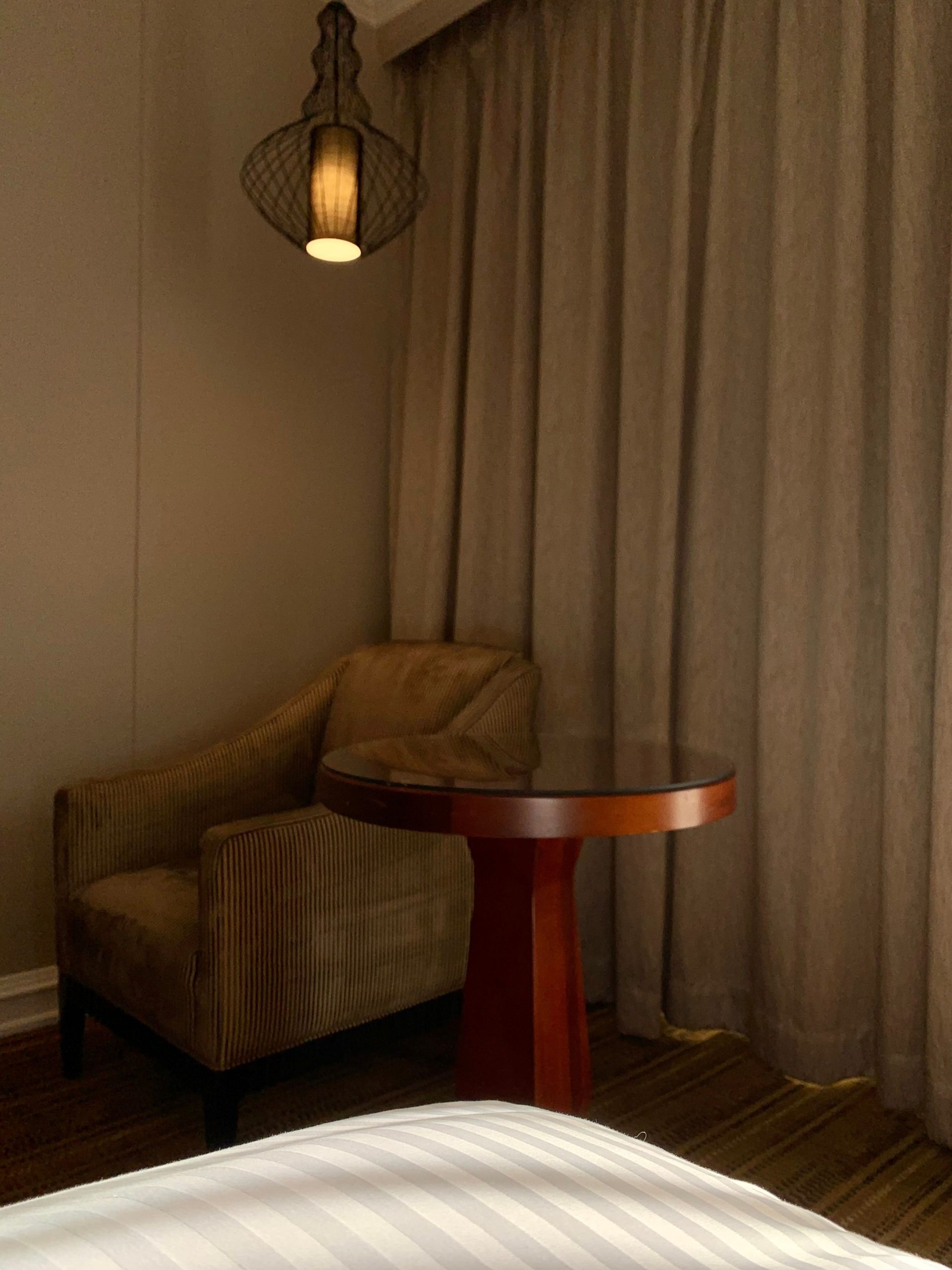 Warm-lit corner of a hotel room featuring an armchair, wooden table, and soft curtains.