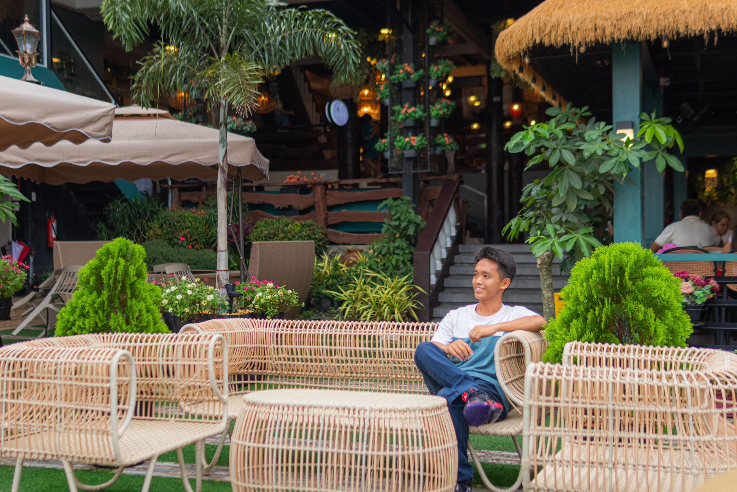 A man relaxing in a tropical garden lounge, surrounded by vibrant plants and wicker furniture.