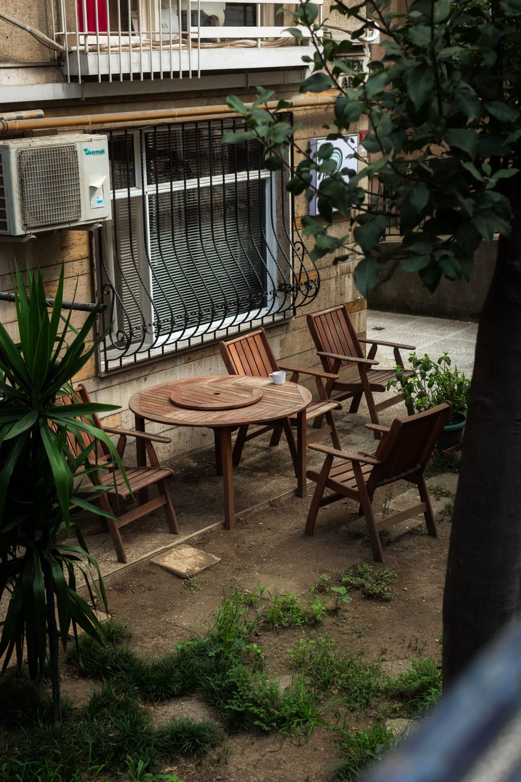 A quaint backyard patio featuring wooden chairs and a round table amidst lush greenery.