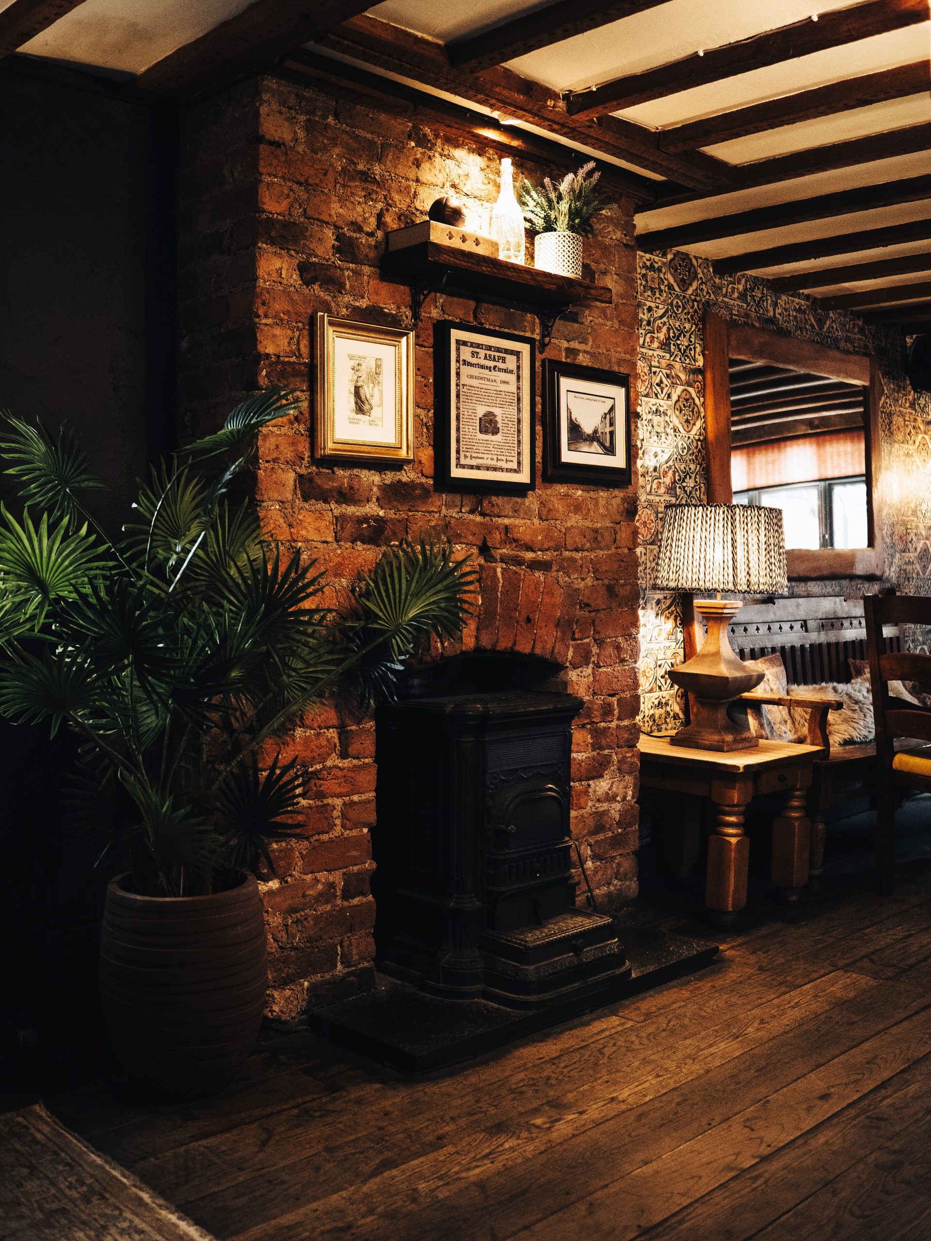 A warm, rustic living room featuring a brick fireplace, wall art, potted plants, and wooden furnishings.