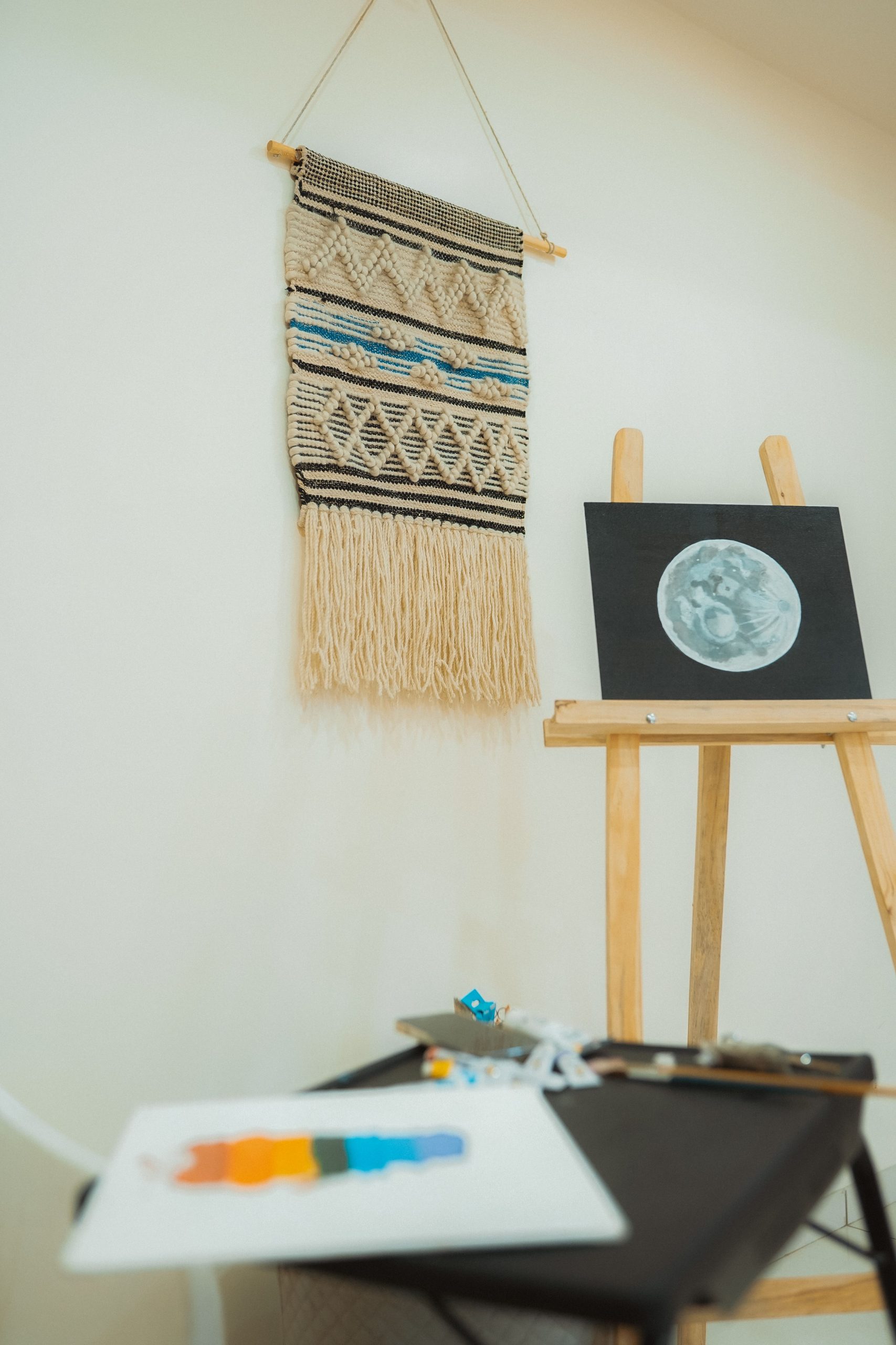 An artist's studio featuring an easel with a moon painting and macrame wall art.