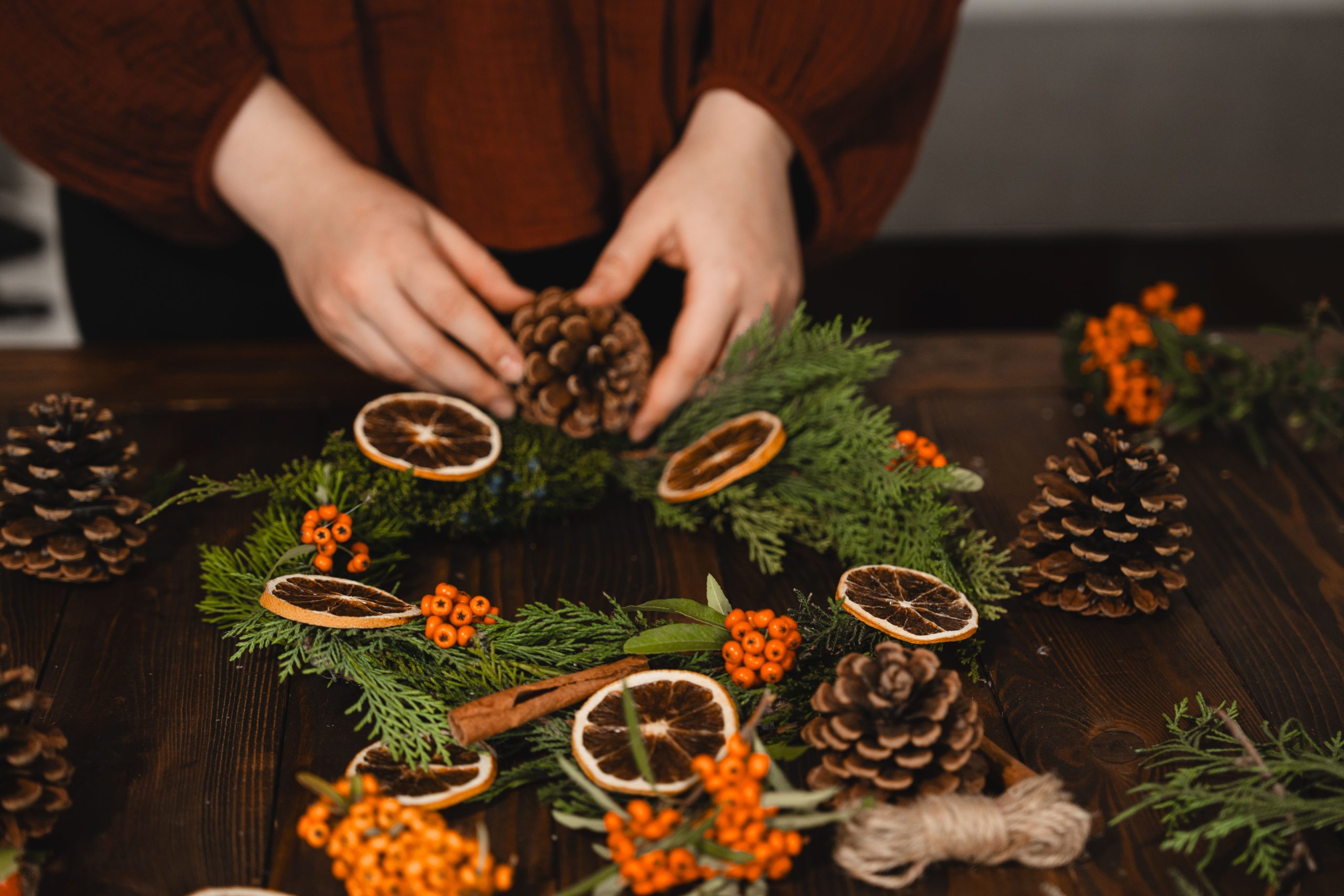 Artisan creating a festive wreath with pinecones and citrus in a cozy setting.