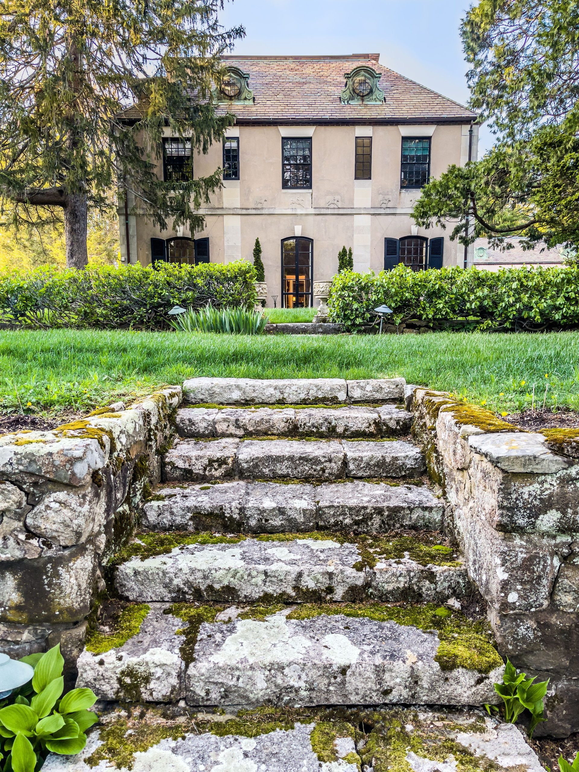 Charming stone steps with moss lead to an elegant historic house surrounded by lush greenery.