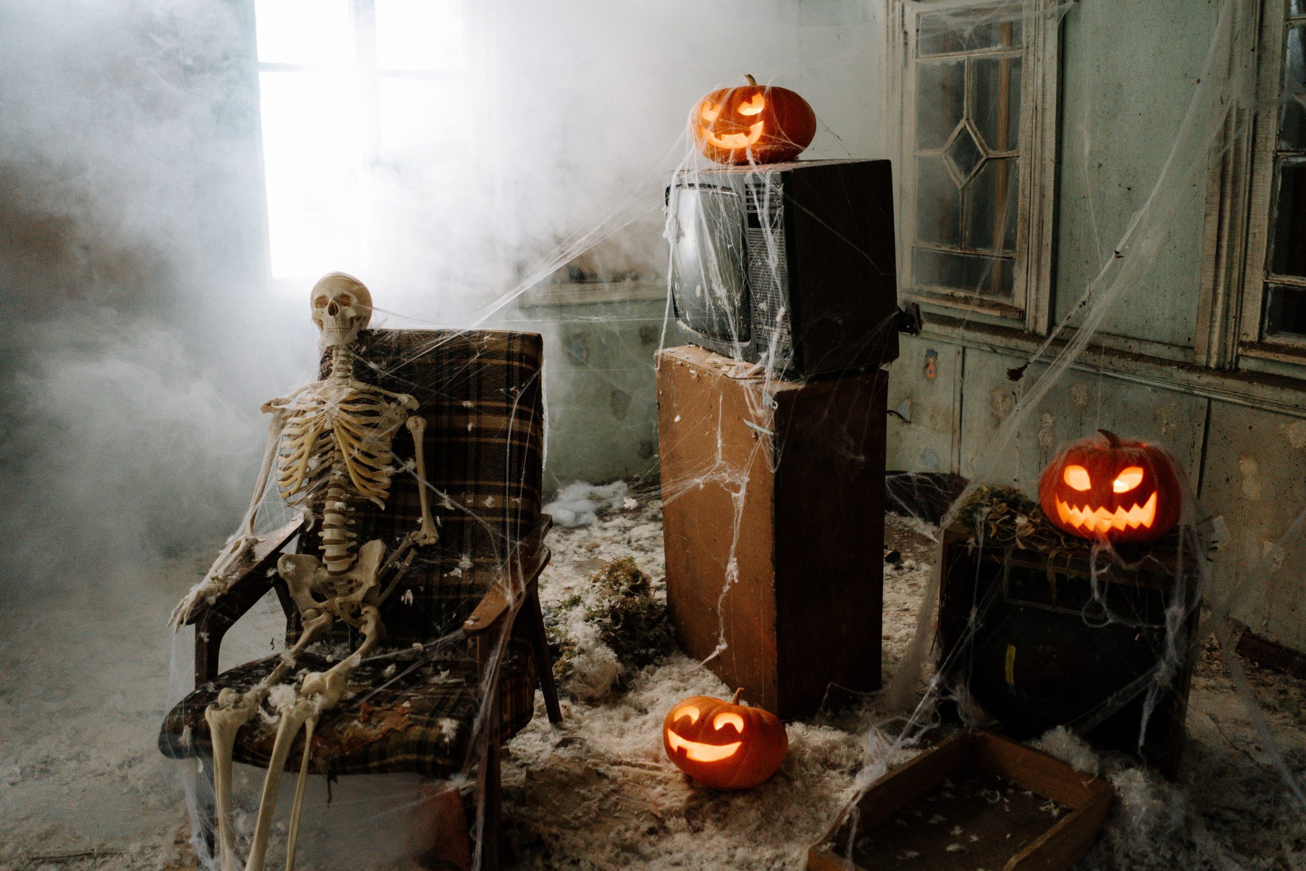 Eerie room with a skeleton, jack-o'-lanterns, cobwebs, and fog for a haunting Halloween vibe.