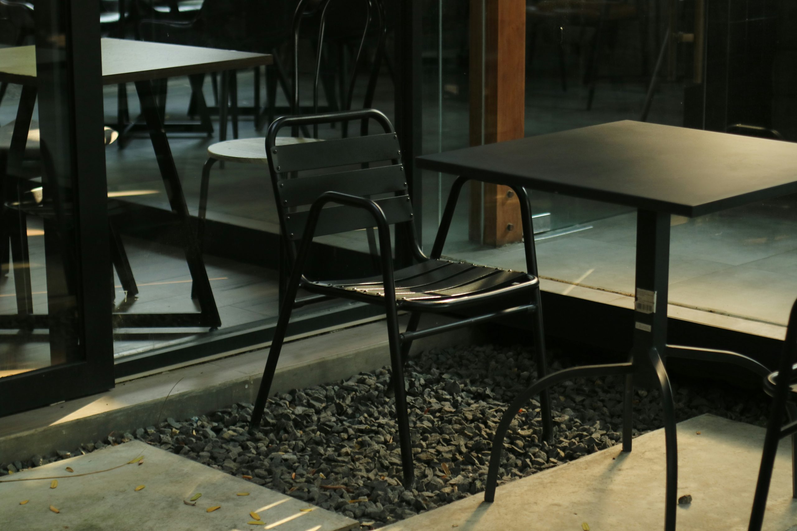 Minimalist outdoor seating area with a black metal chair and table, surrounded by gravel and glass walls—ideal for urban lifestyle, café, and design concepts
