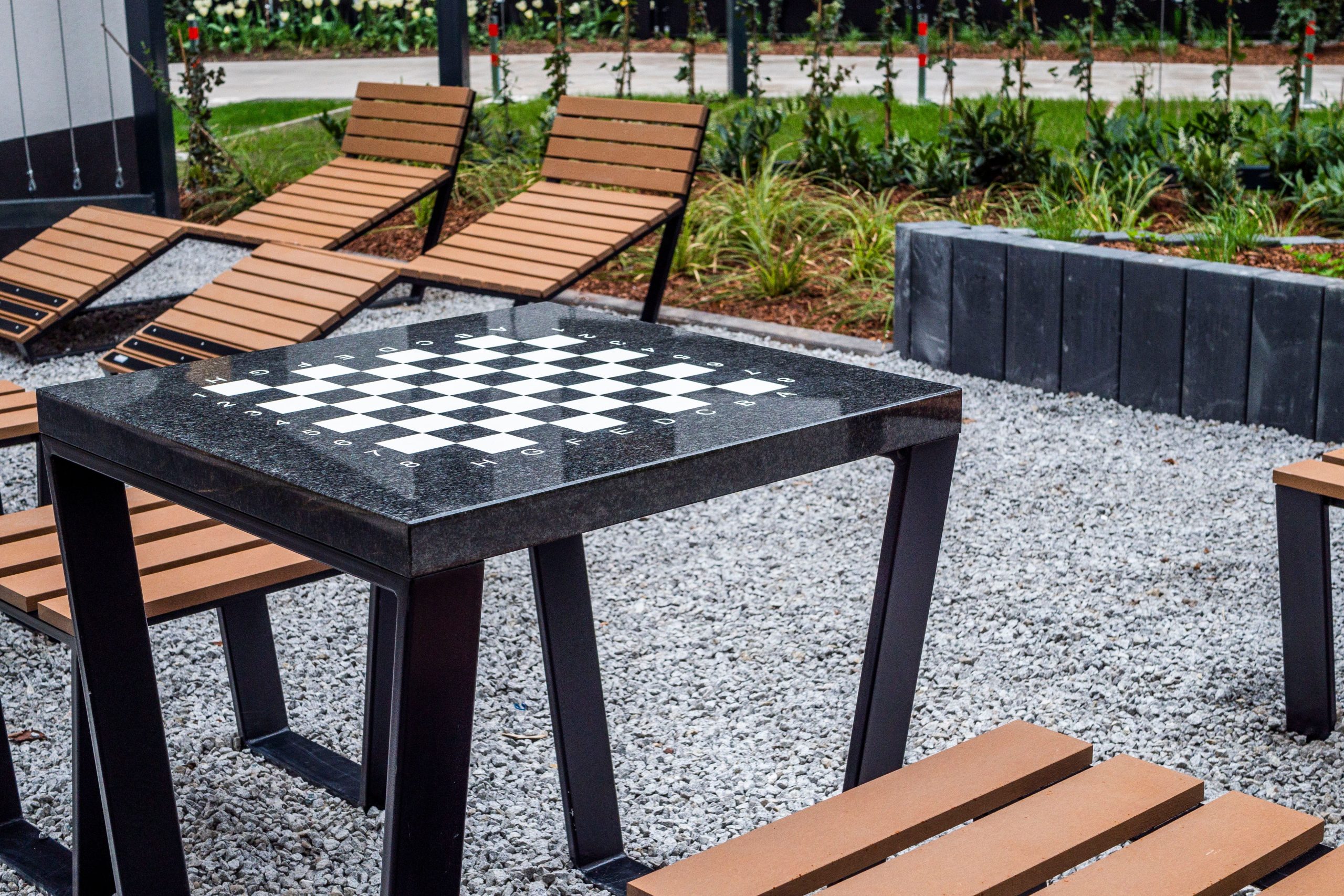 Modern outdoor chess table area with wooden lounge chairs on gravel.