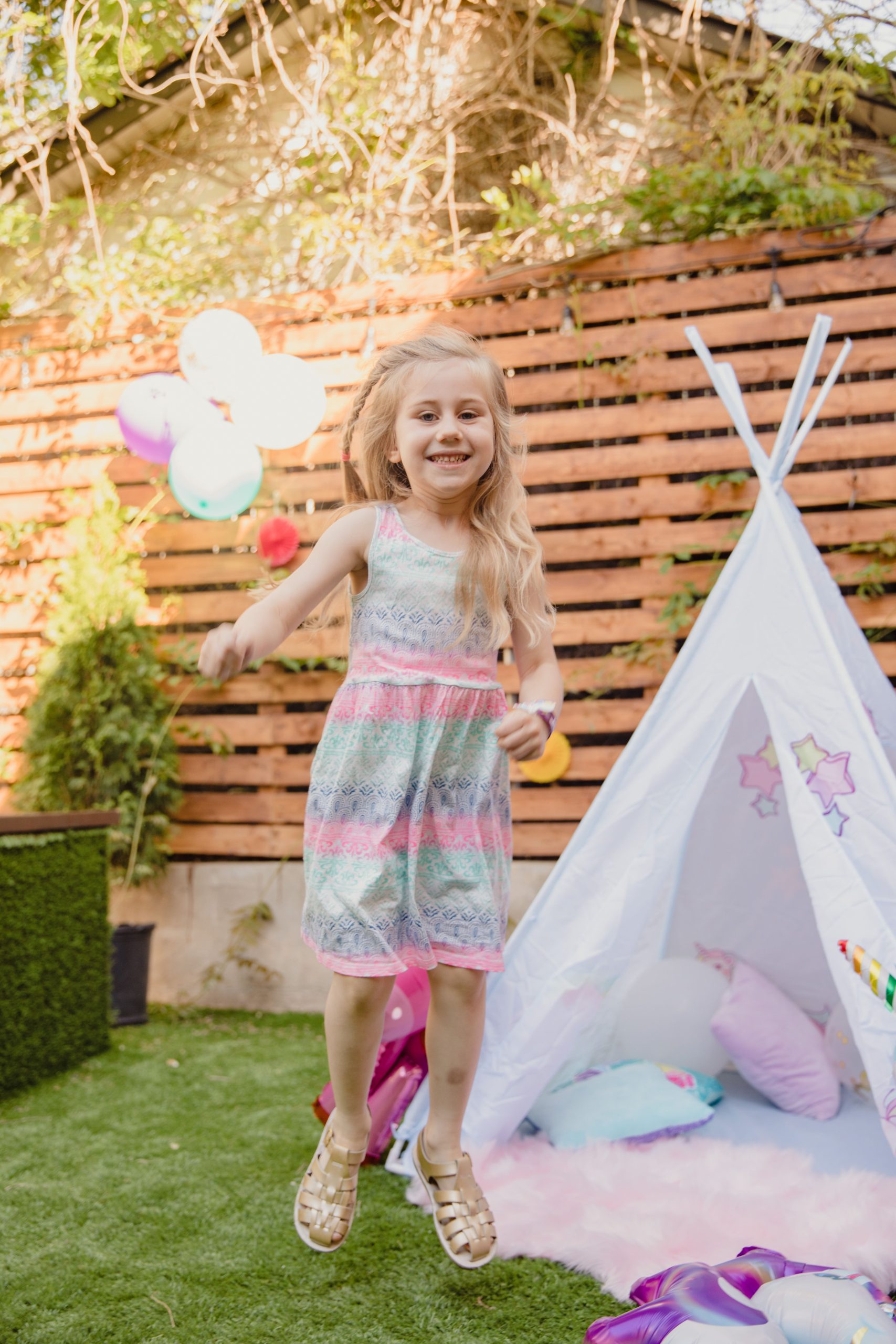 Smiling girl jumping in a playful backyard with a teepee and balloons, capturing summer fun.