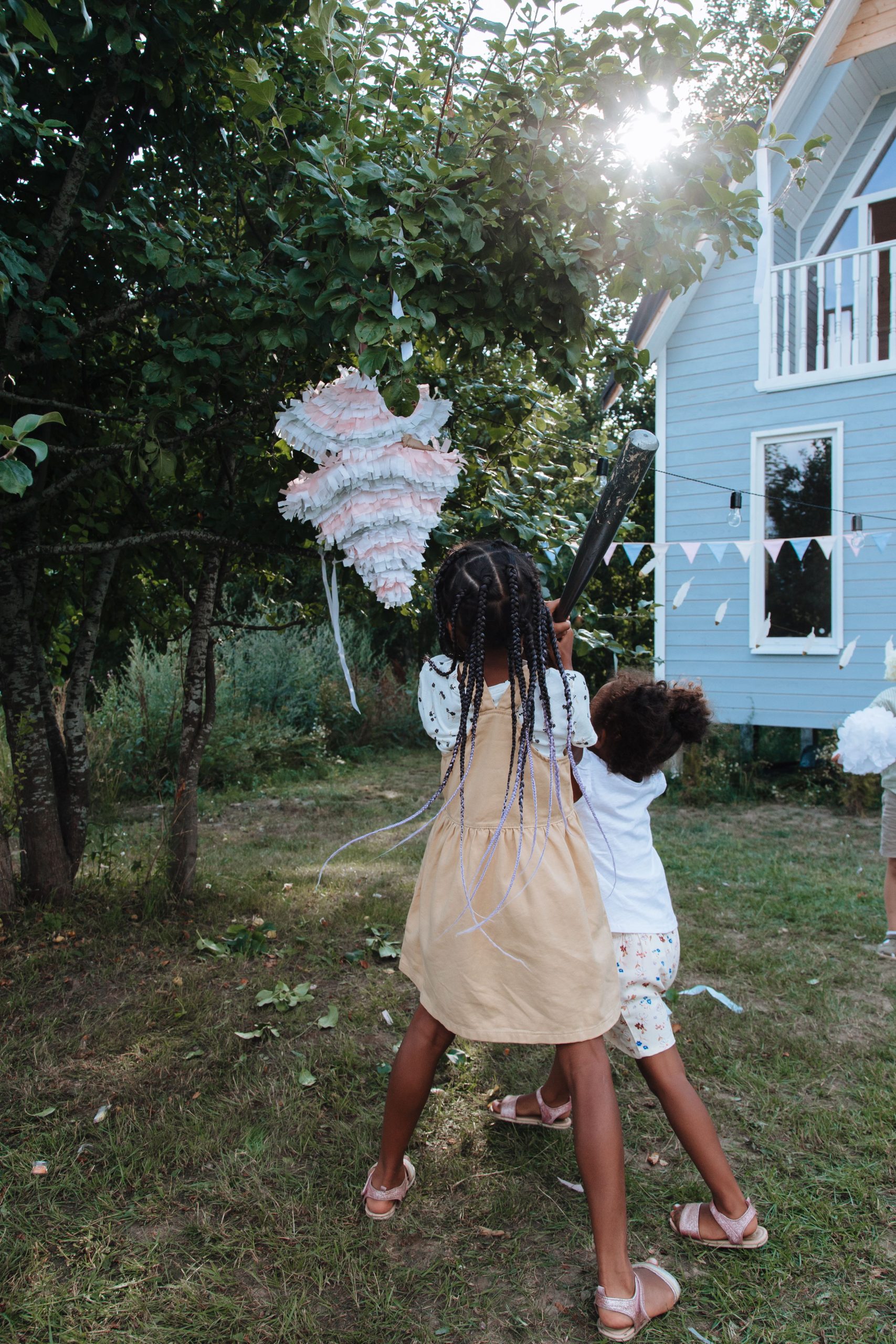 Two children hitting a pinata in a garden setting with trees and a house.