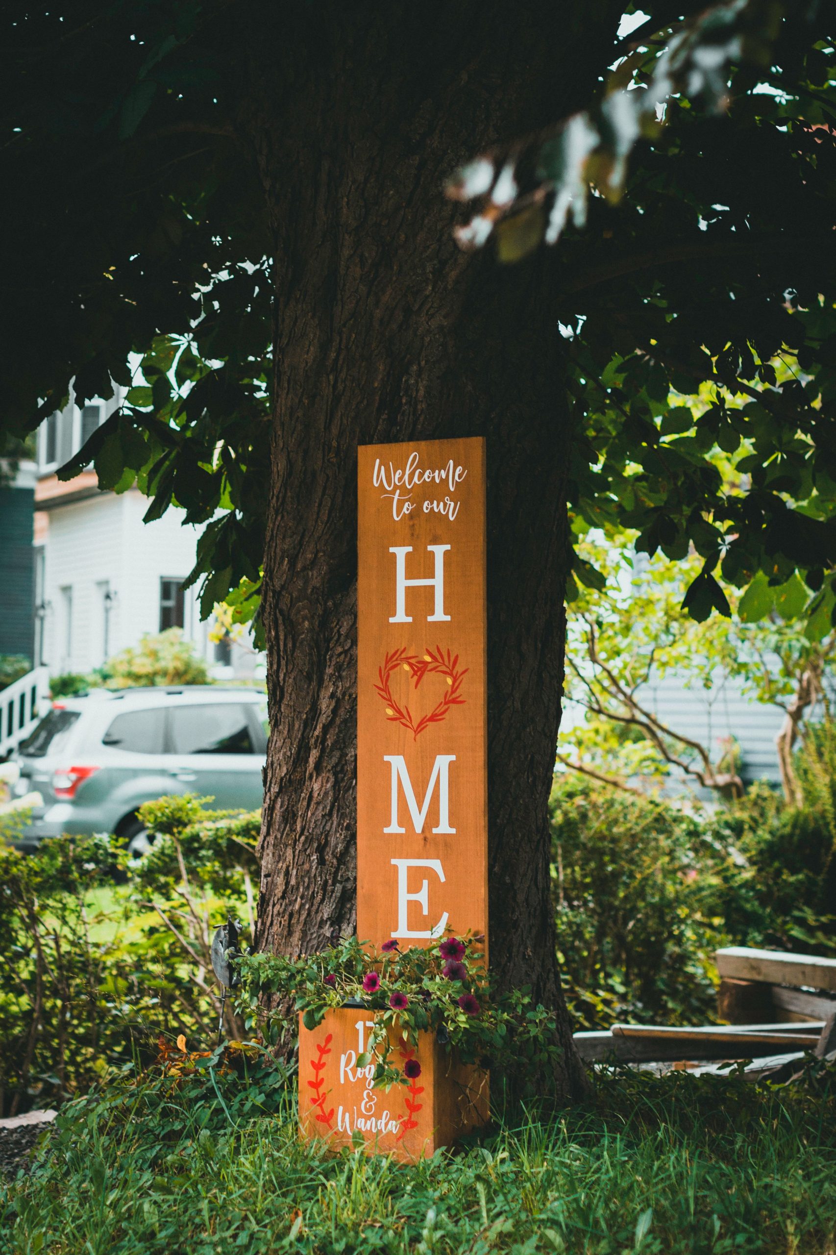 Vertical wooden sign saying 'Welcome to our Home' under a tree outdoors.