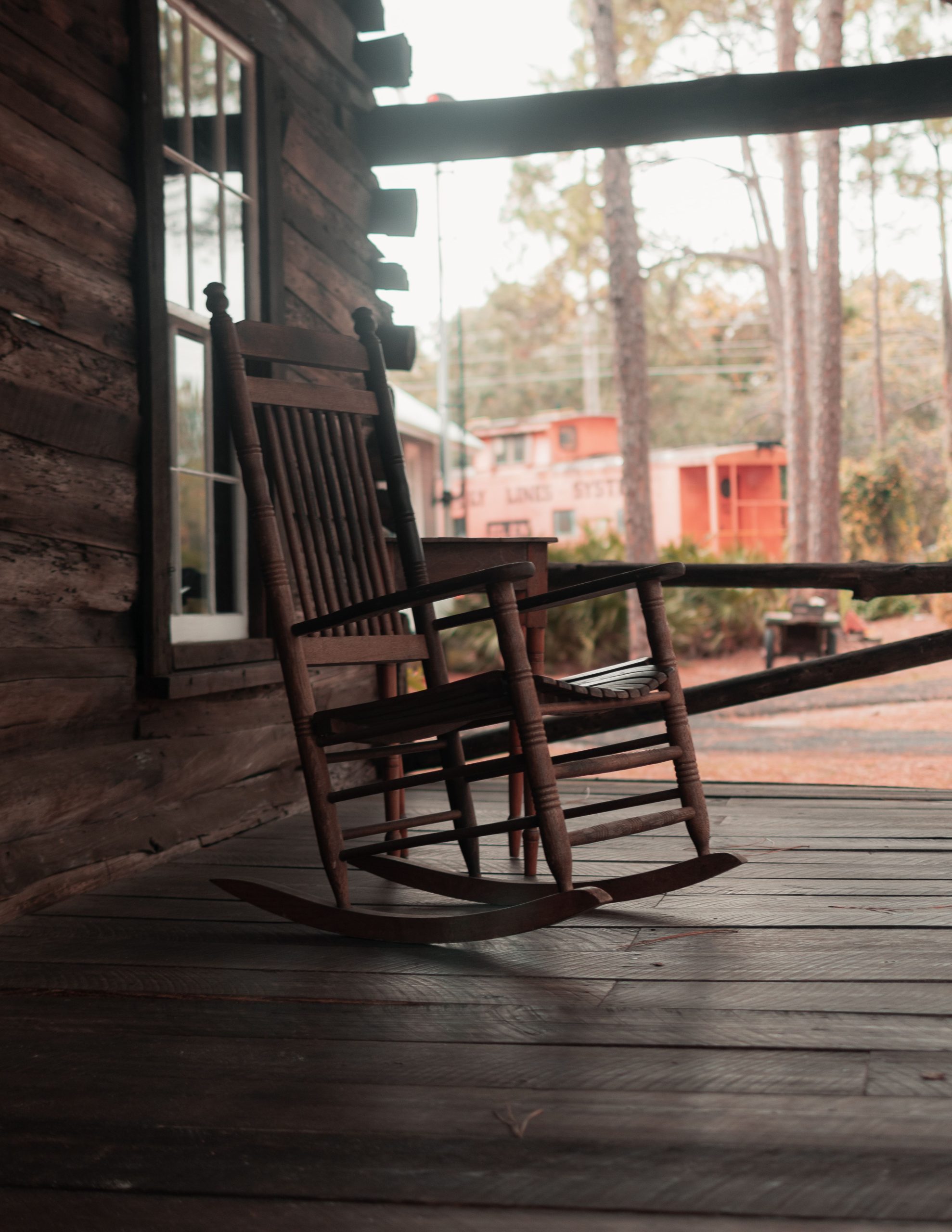 Wooden rocking chair located on terrace of wooden house with big window against wooden floor and trees and building in daytime