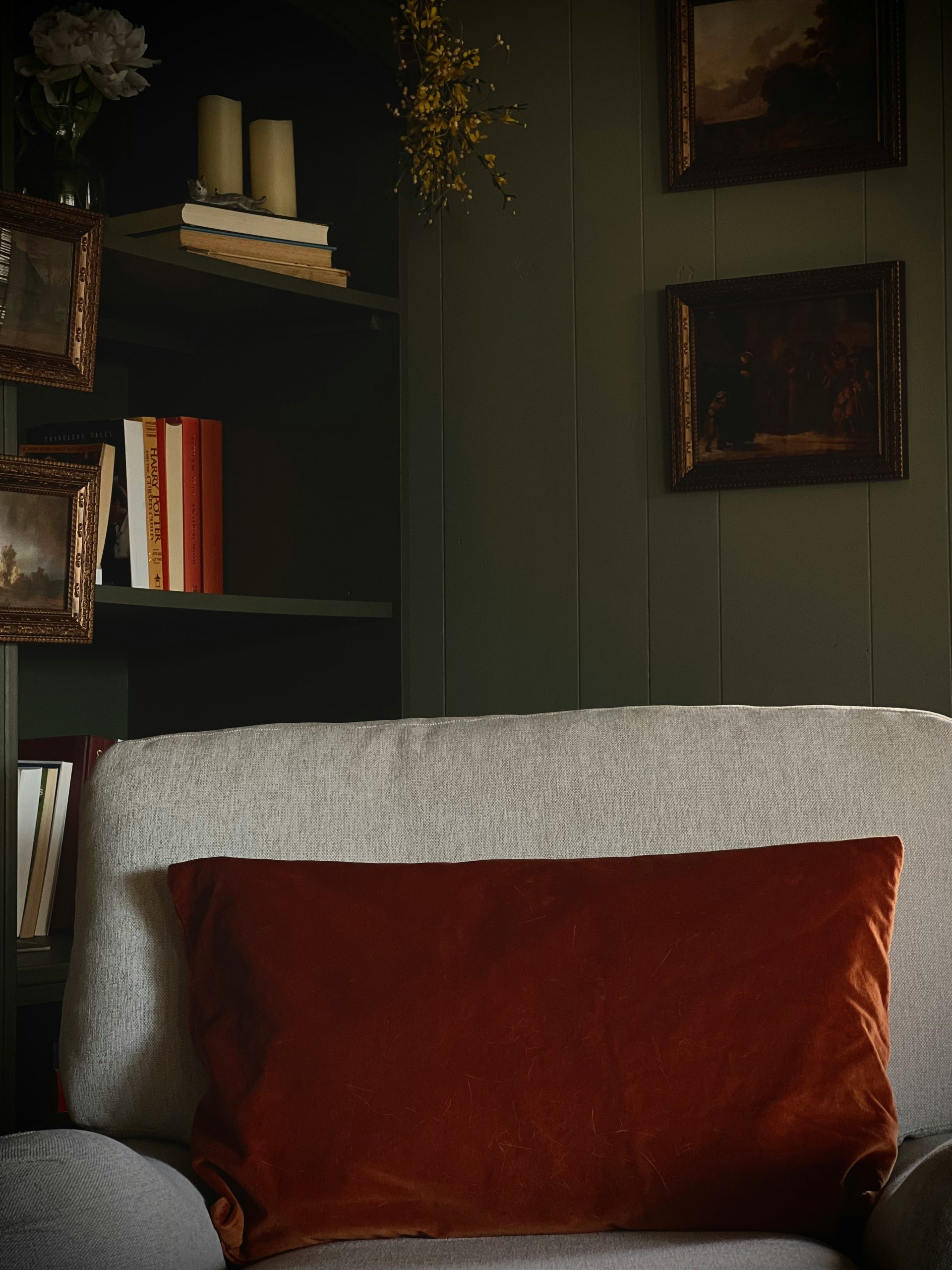 A close up of a dark, cozy reading corner. Antique bronze framed art prints sit on the walls and bookshelf. An off white colored English armchair looks inviting, ready for anyone to curl up with a goo