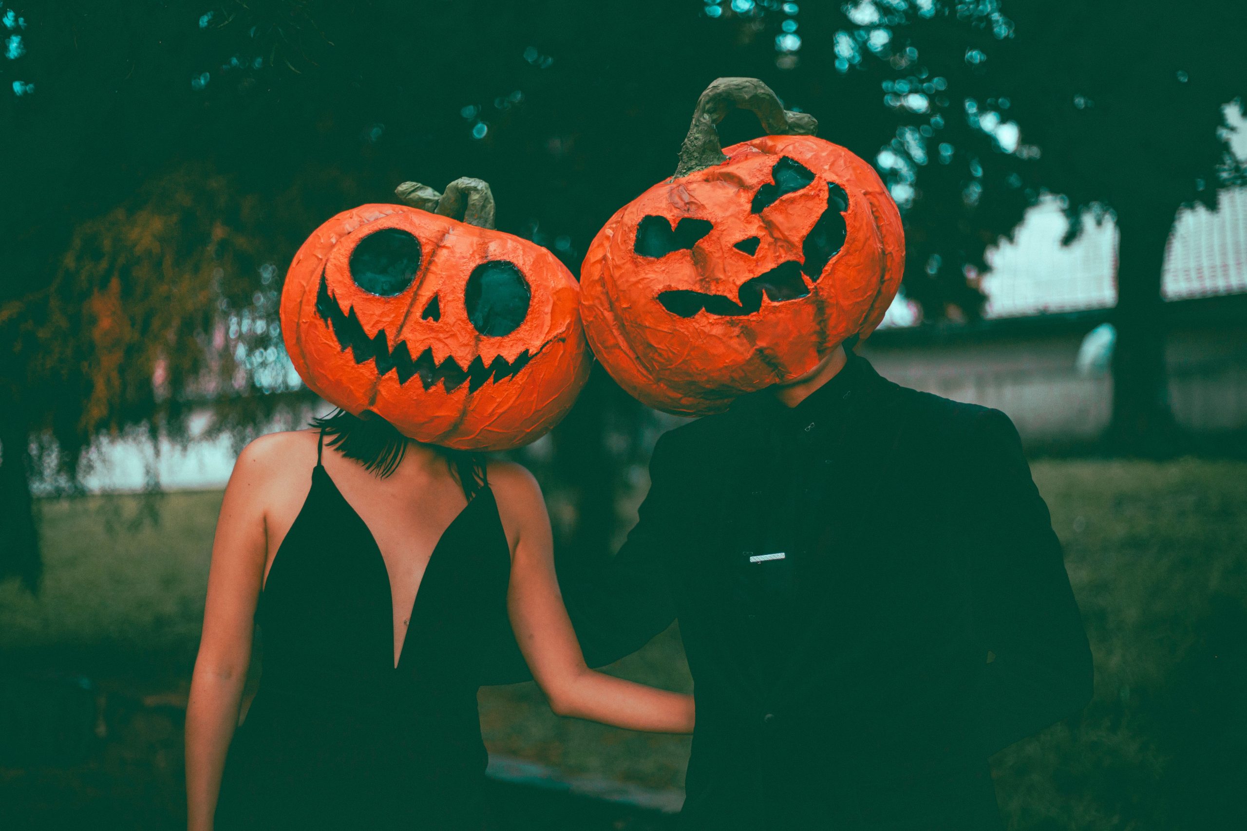A couple in stylish Halloween costumes with pumpkin masks, posing outdoors during fall.