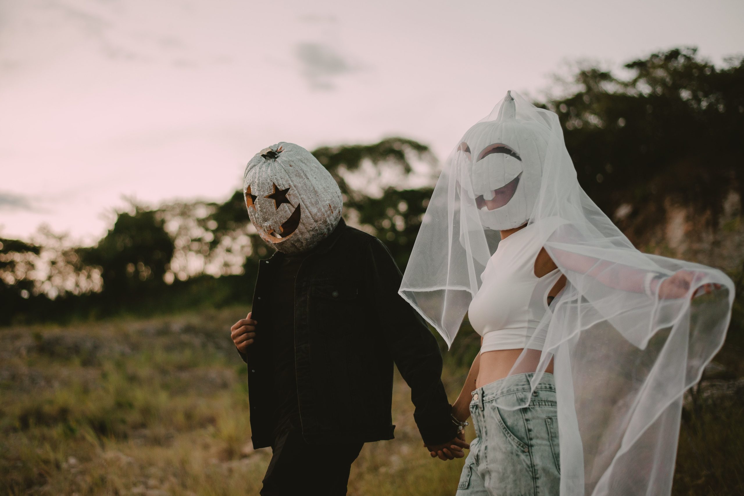 A couple wearing pumpkin head costumes and veils walking hand in hand in a rural setting.