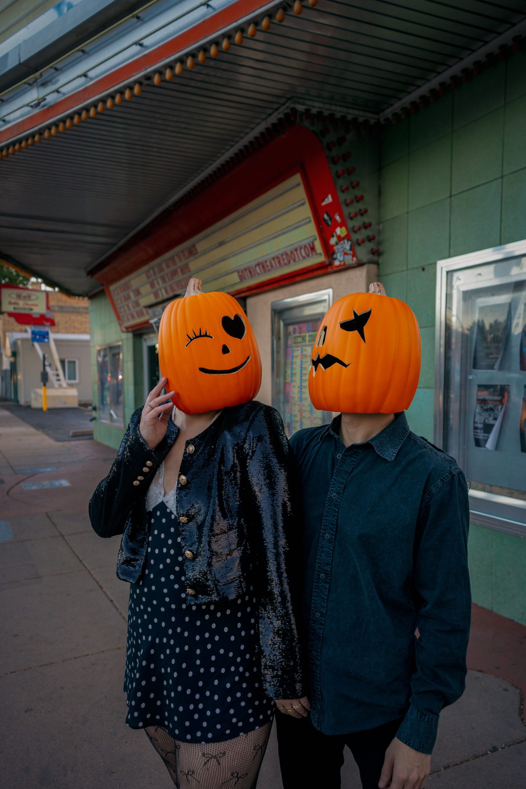 A couple with carved pumpkin heads poses outside a theater in Englewood, capturing fall vibes.