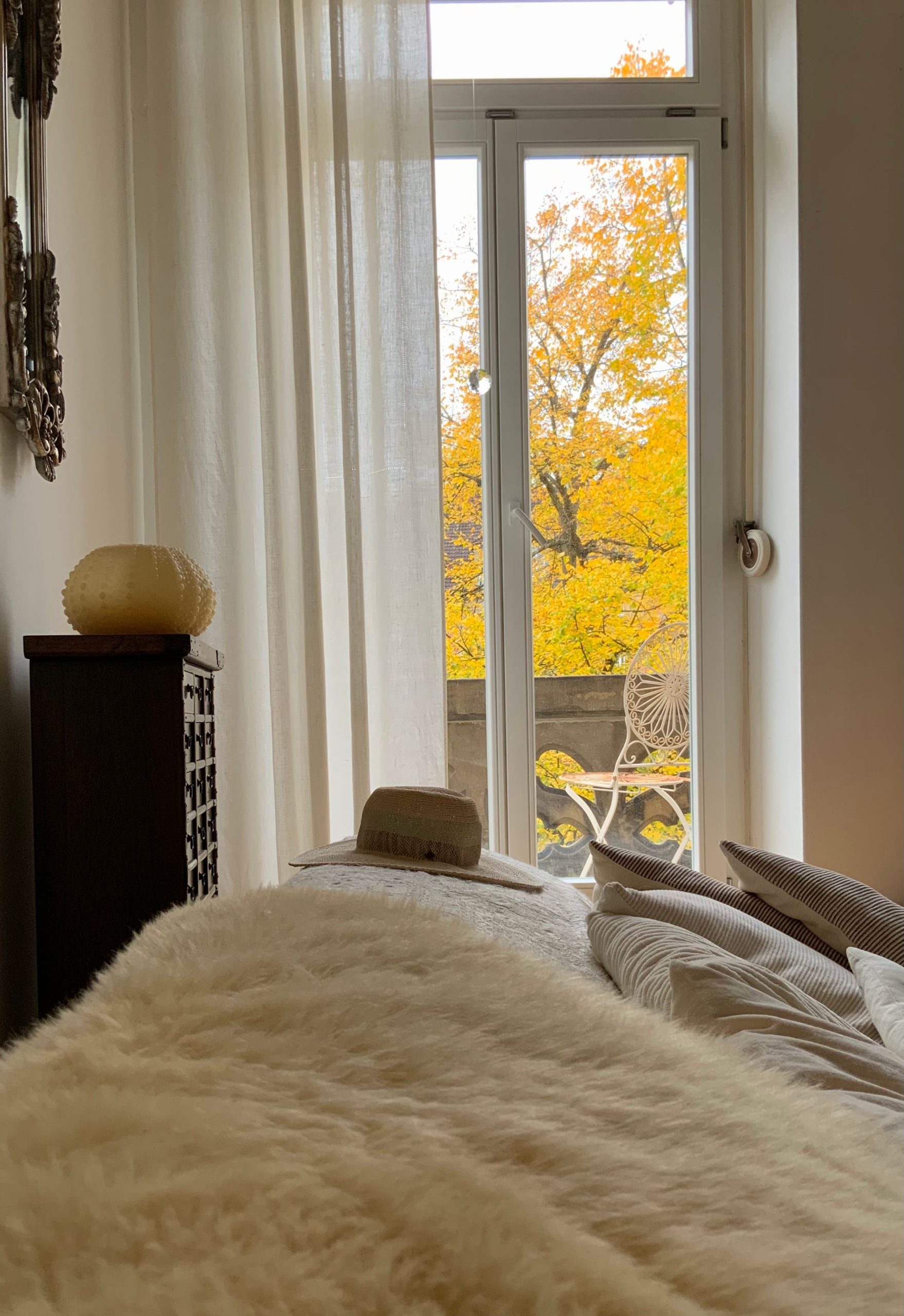 A cozy bedroom with a fluffy bed overlooking vibrant autumn foliage through a tall window.