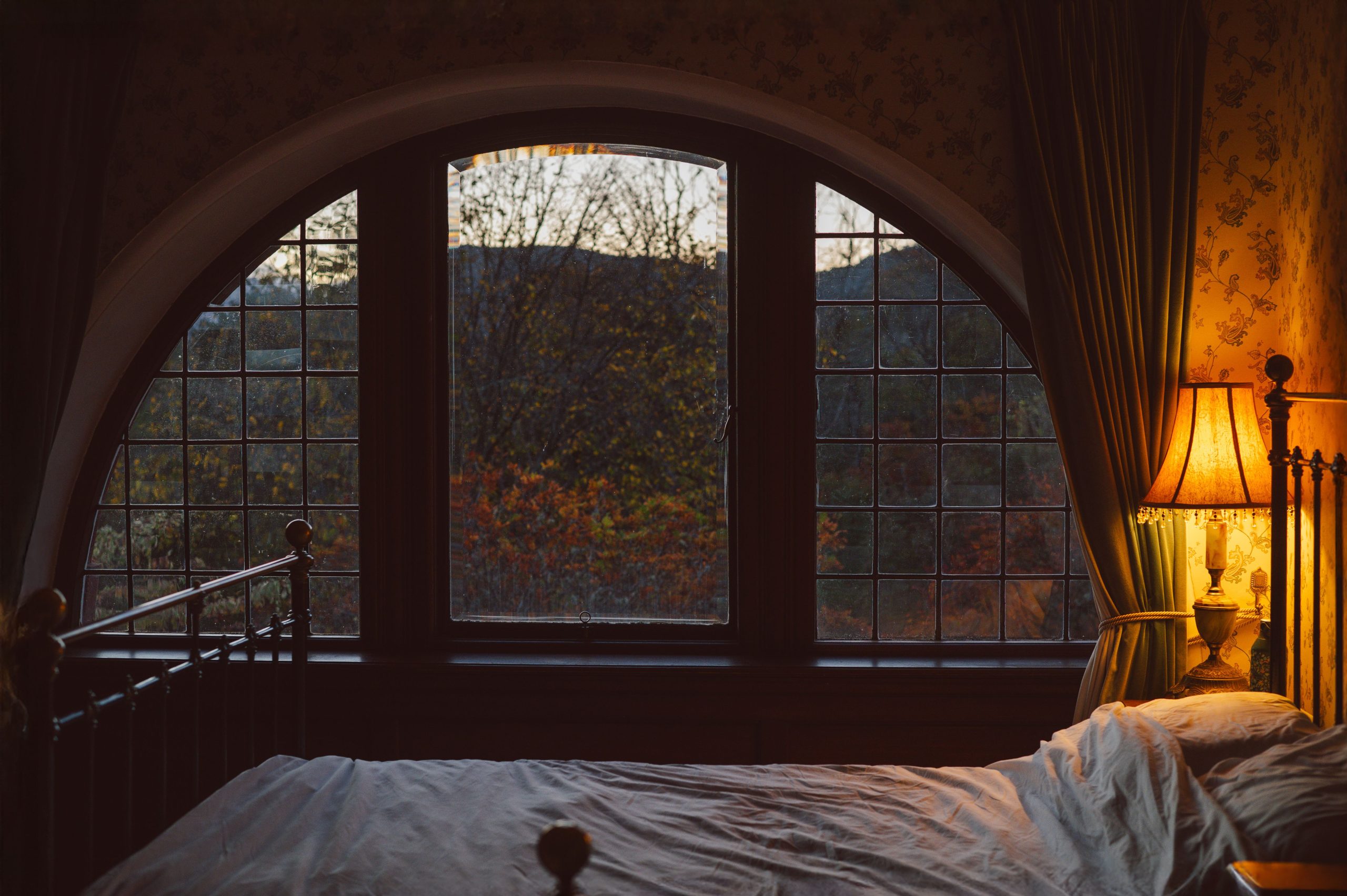 A cozy Victorian-style bedroom with a large arched window and soft lighting from a bedside lamp.