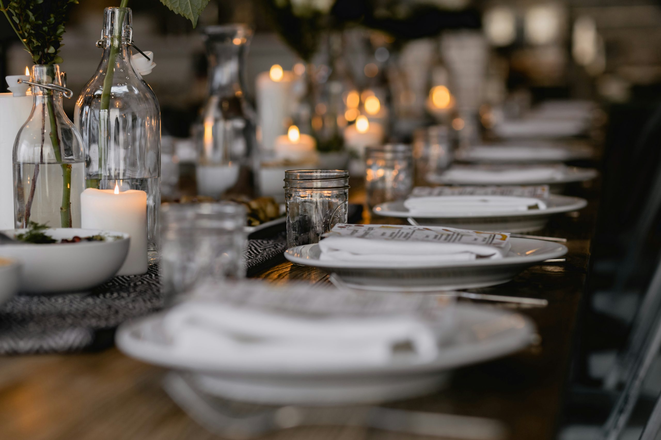 A long dining table set with white plates, candles, and glass bottles