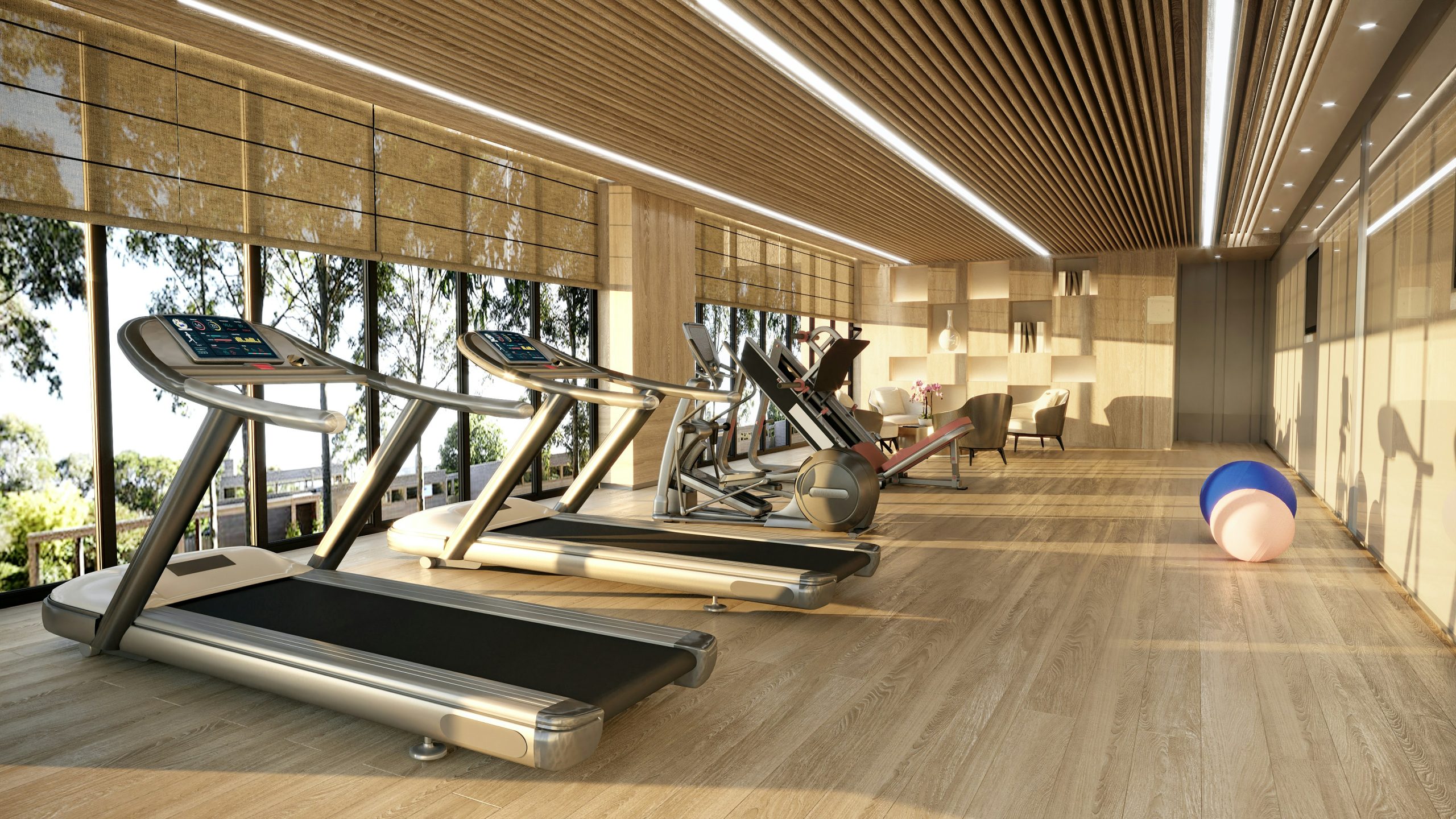 A modern residential gym with sleek wood flooring and a slatted ceiling. The space features treadmills, exercise machines, and a lounge area, with large windows overlooking a green, sunlit landscape.