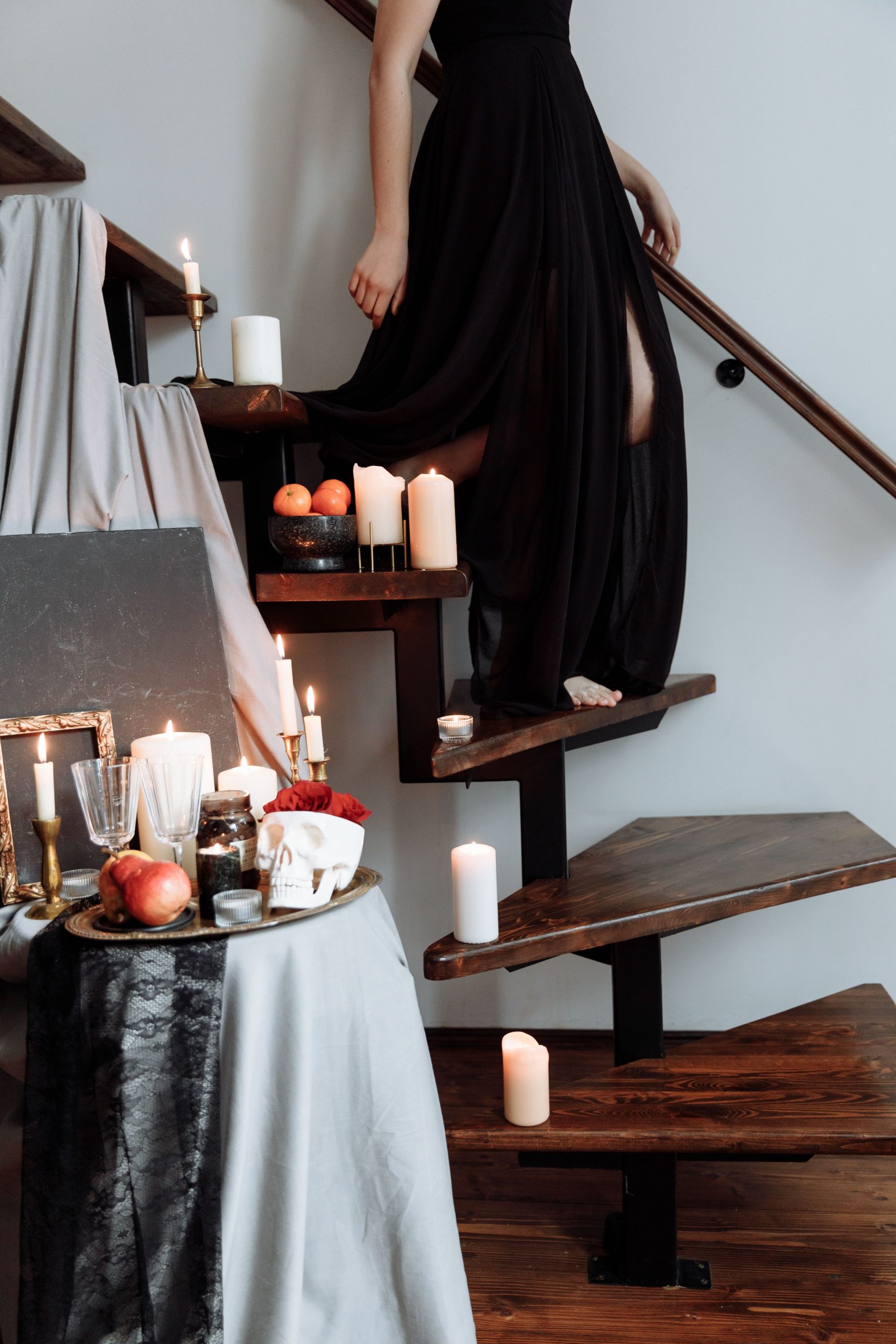A person descends wooden stairs adorned with candles and elegant decor, wearing a flowing black dress.