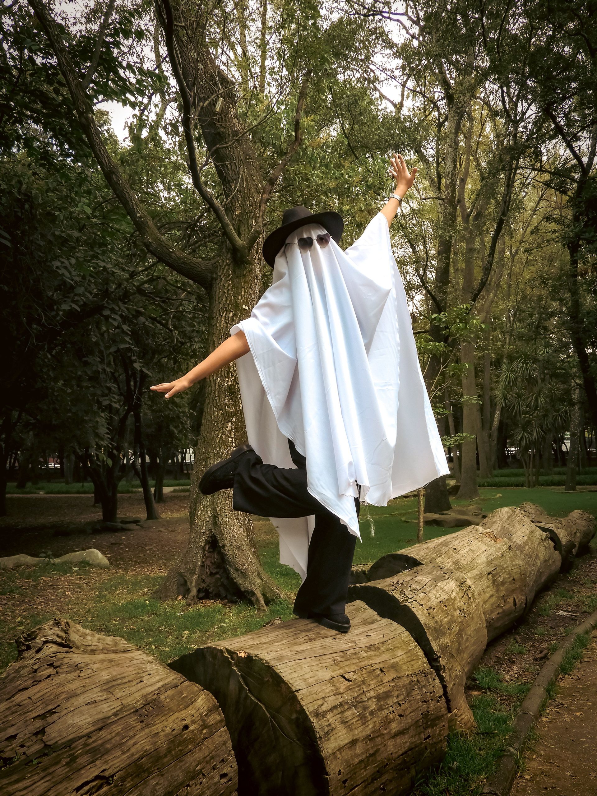 A person in a ghost costume balancing on a log in a park setting, showcasing Halloween fun.