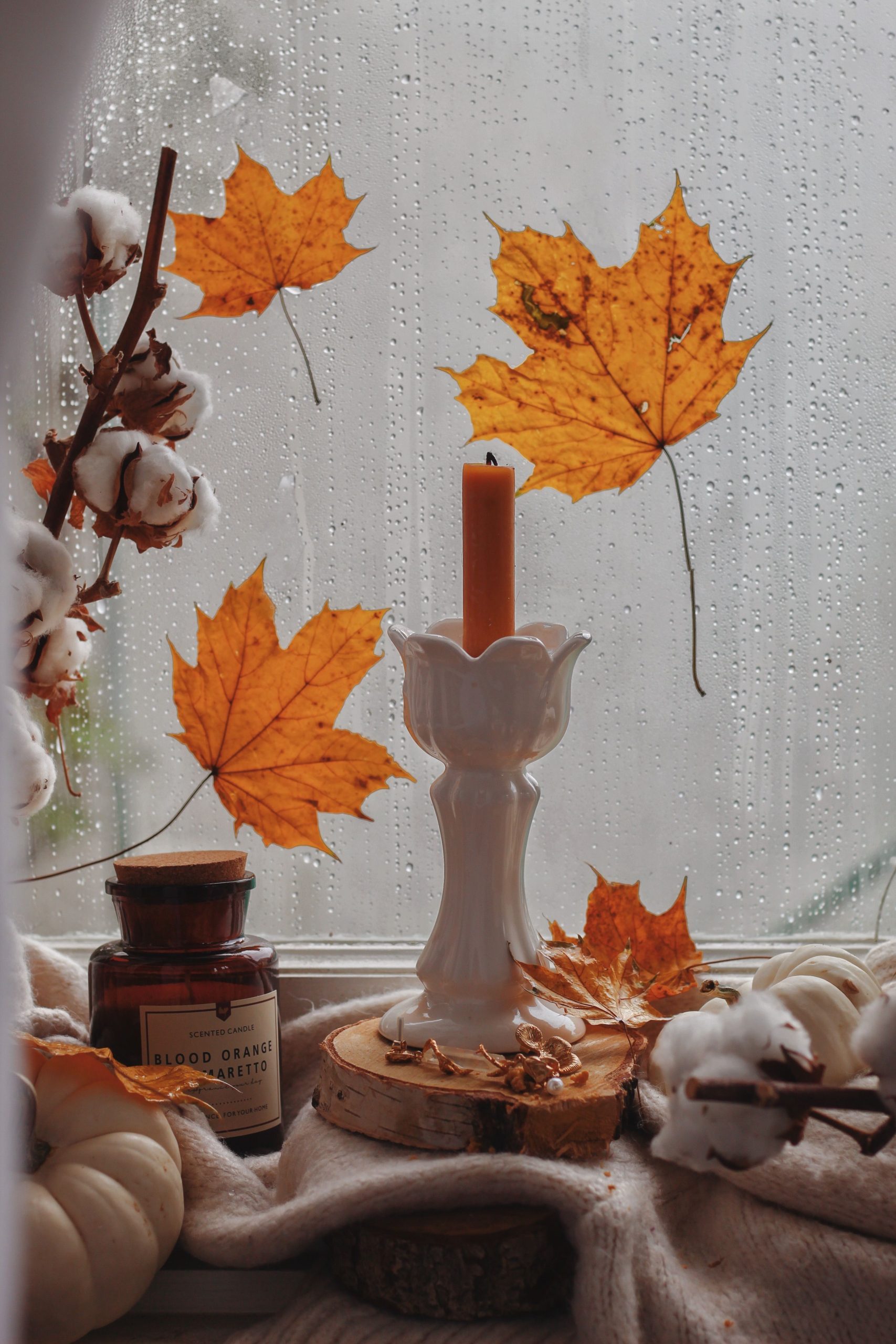 A serene autumn scene with candlelight, maple leaves, and cozy decor by a rainy window.