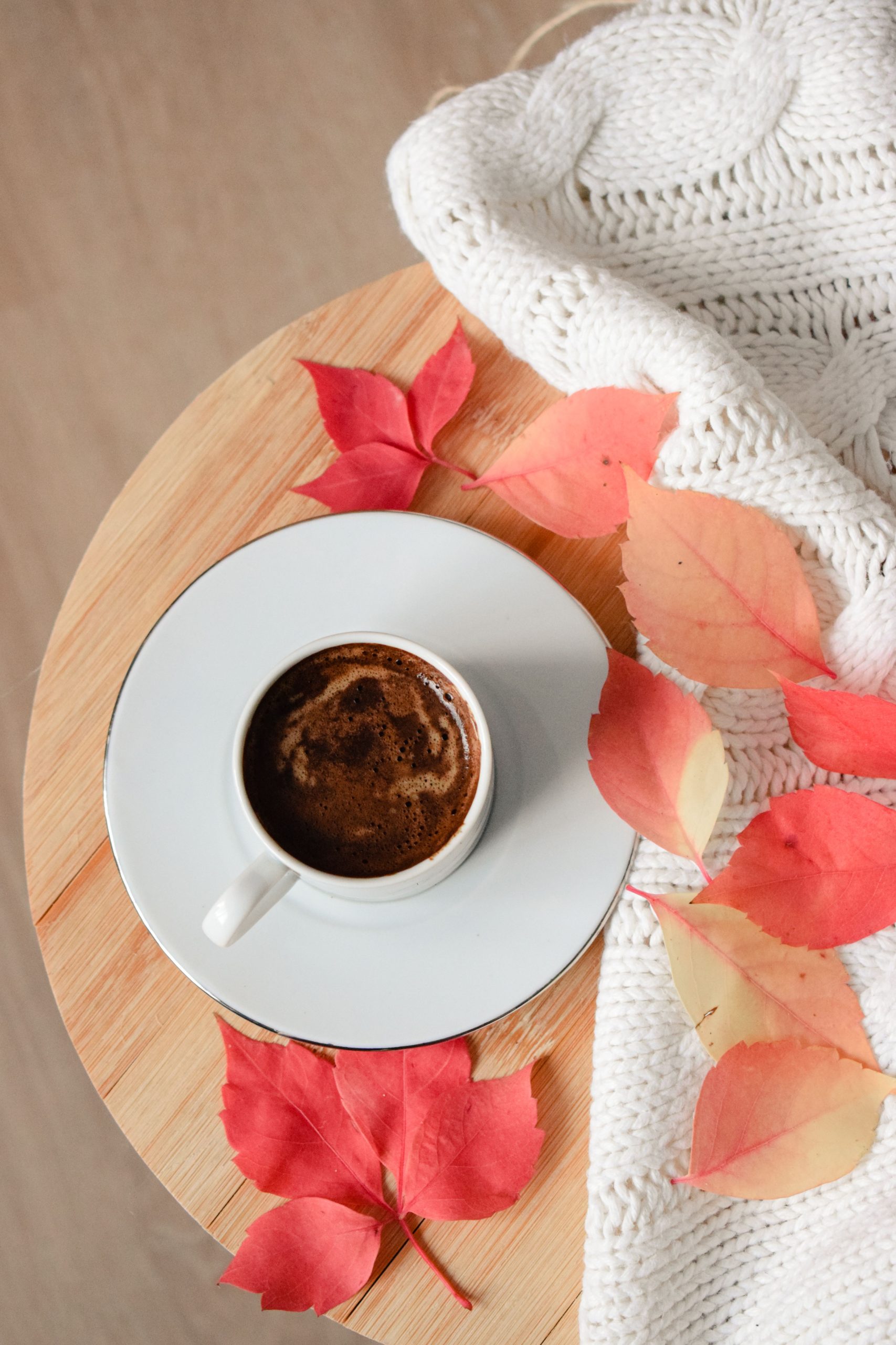 A warm cup of Turkish coffee surrounded by autumn leaves and a cozy knit blanket on a wooden table.