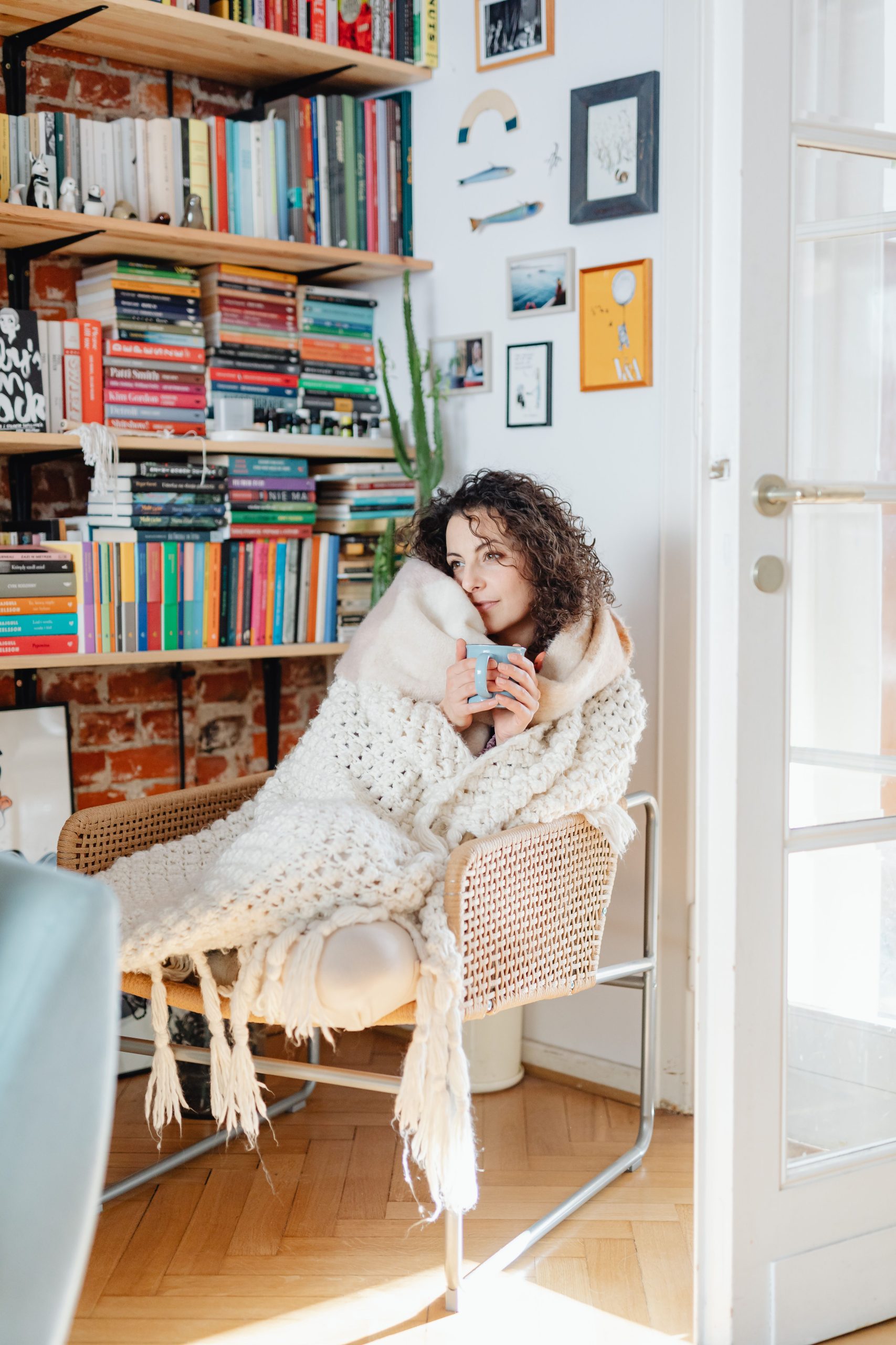 A woman wrapped in a blanket enjoying a warm drink in a cozy room filled with bookshelves.