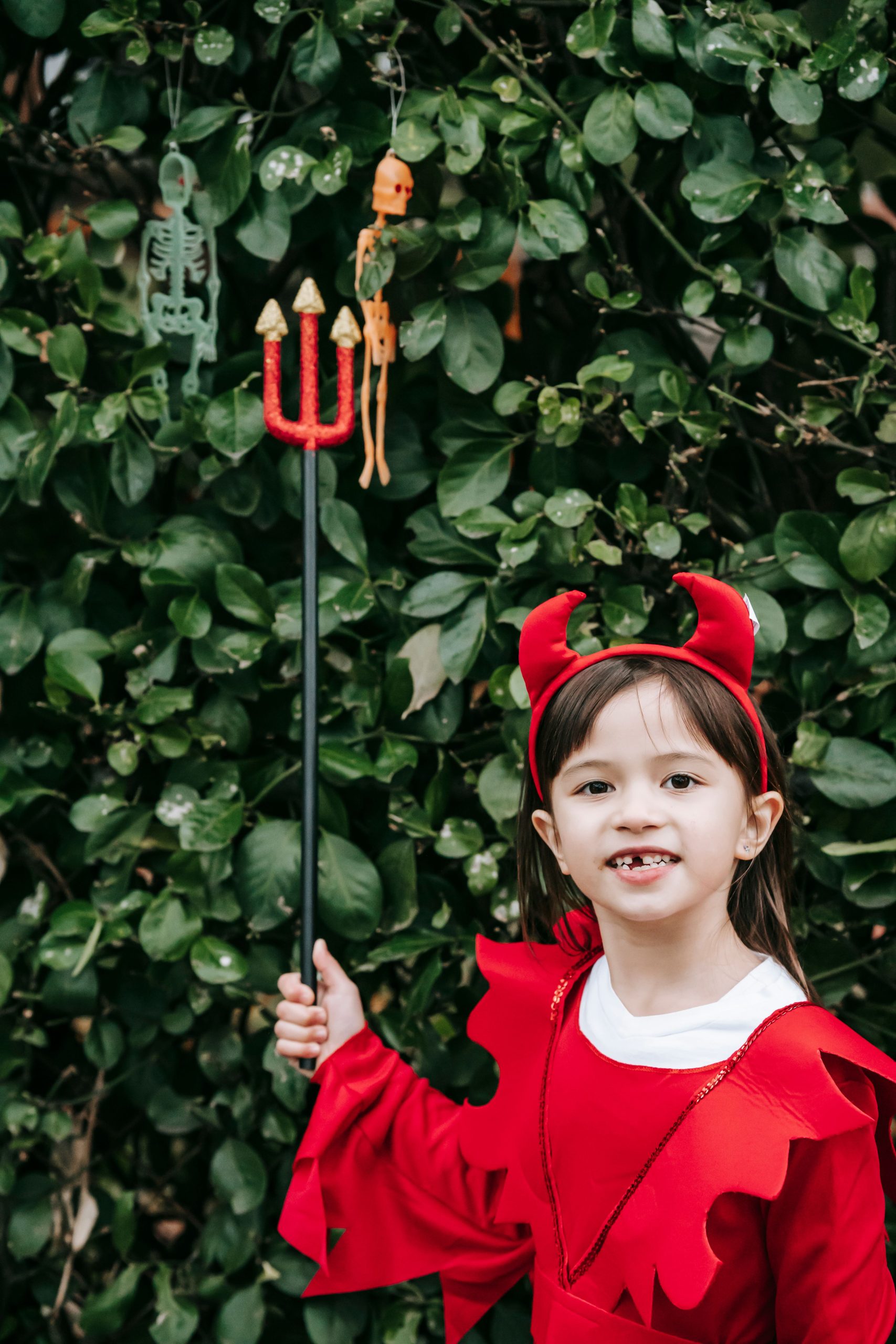 Adorable child in headband with devil horns standing near bush and holding trident while looking at camera