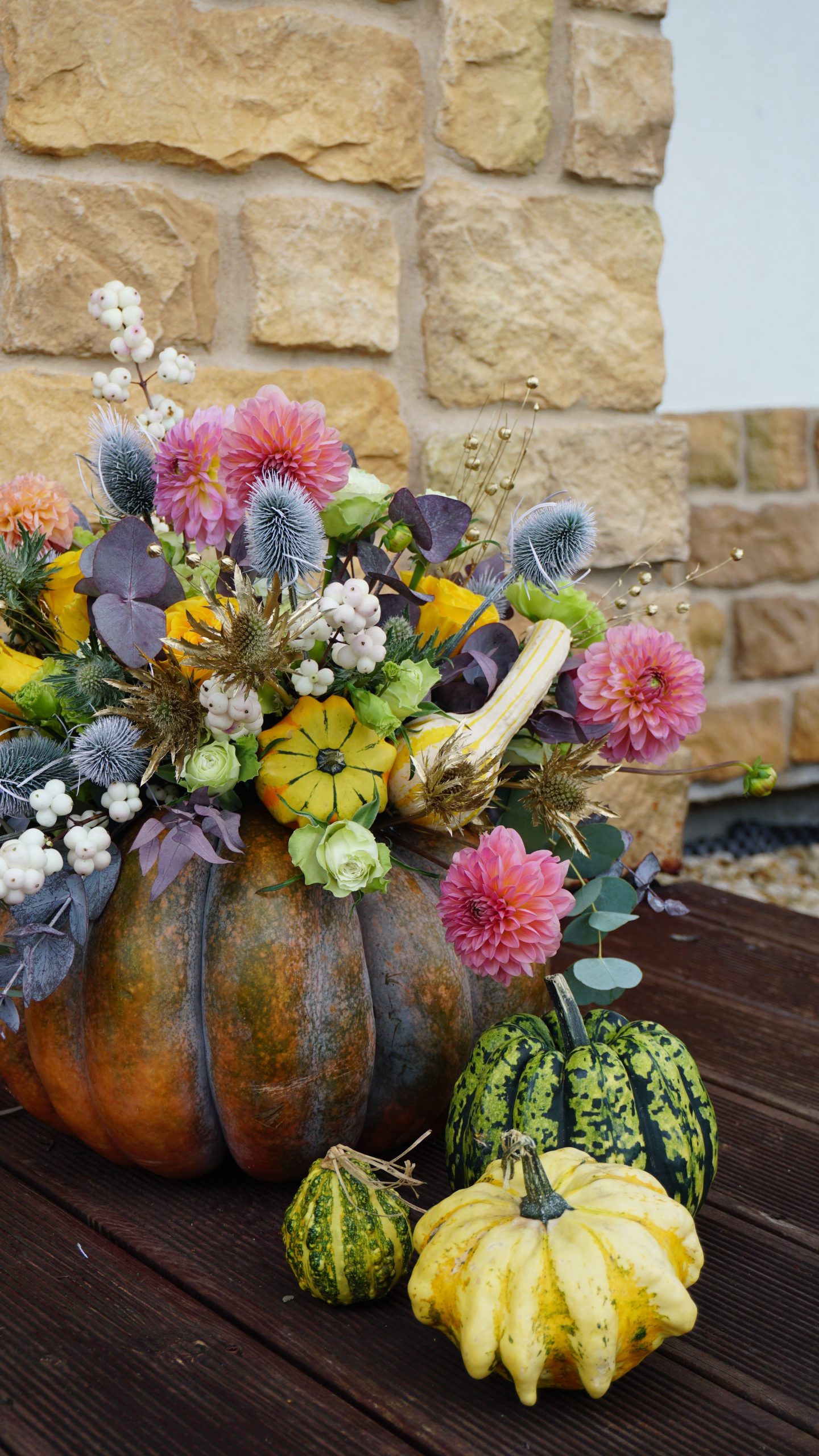 Autumn floral arrangement in a pumpkin with vibrant flowers and gourds against a stone wall backdrop.