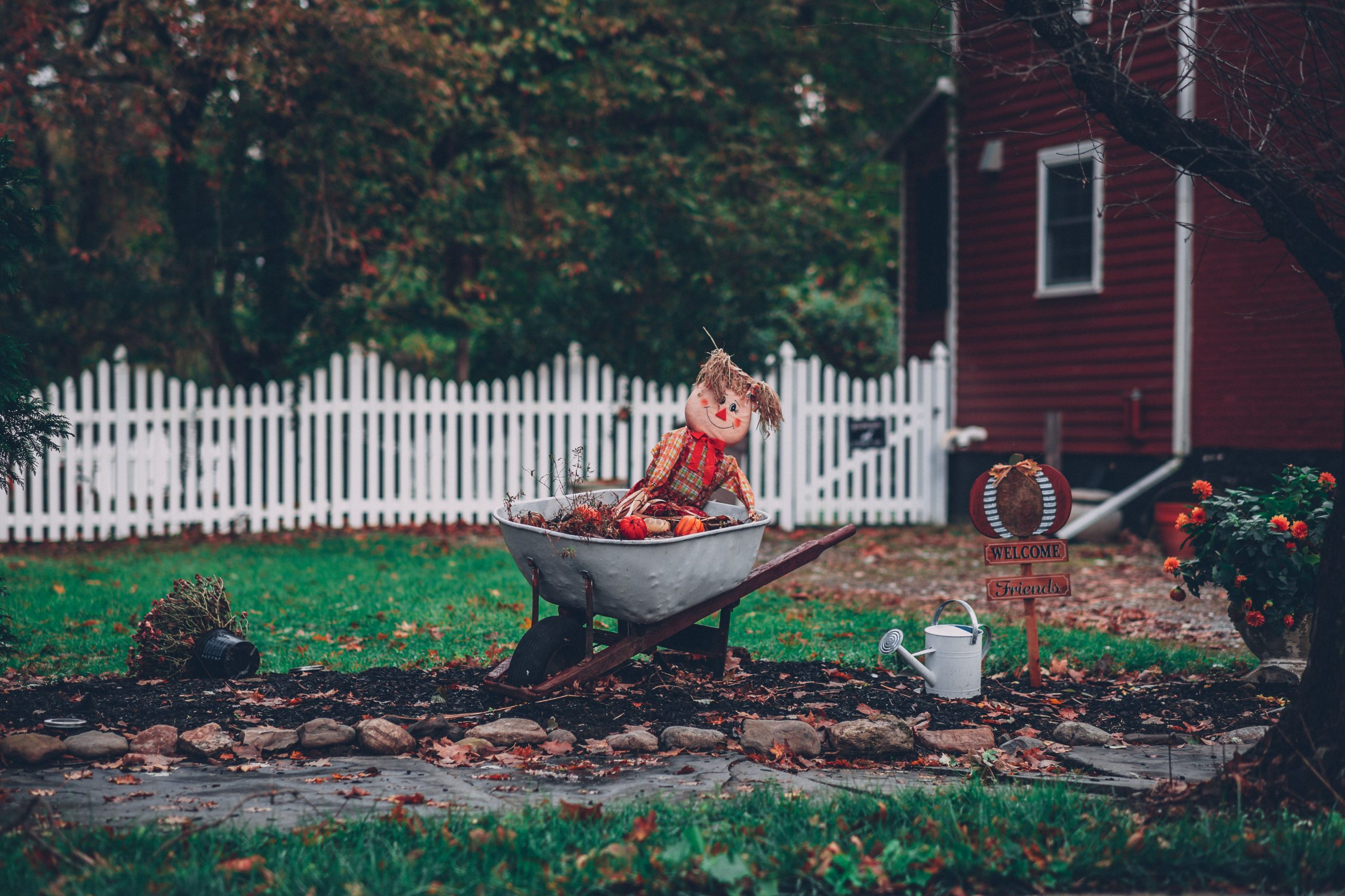 Charming autumn garden with a scarecrow in a wheelbarrow, surrounded by vibrant foliage and a wooden house.