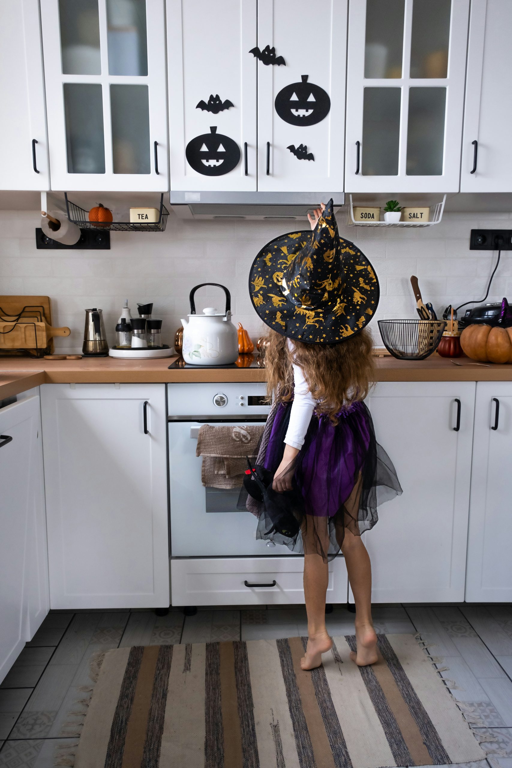 Child decorates the kitchen in home for Halloween. Girl in a witch costume plays with the decor for the holiday - bats, jack lantern, pumpkins. Autumn comfort in house, Scandi-style kitchen, loft
