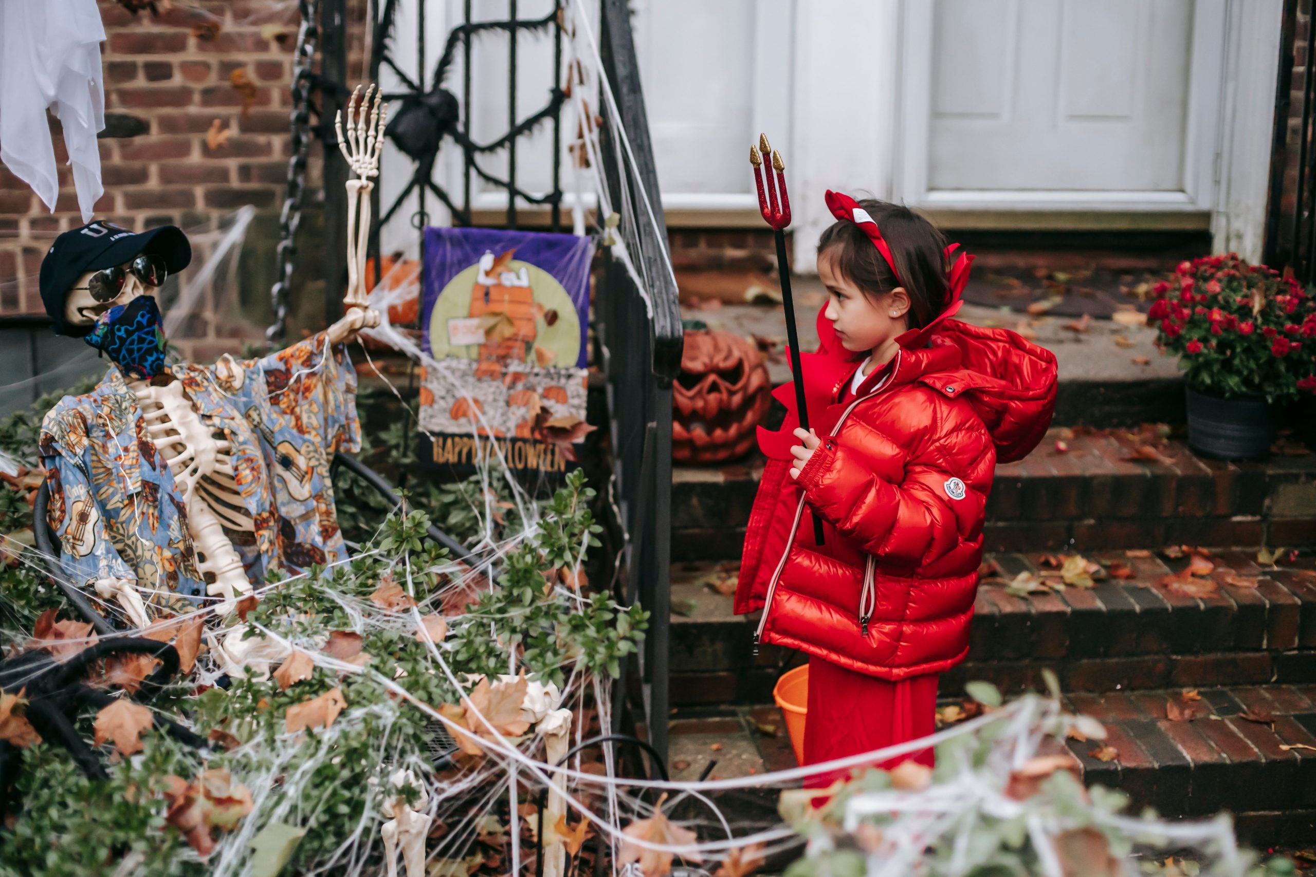 Child in red devil costume with horns and pitchfork standing with bucket near skeleton and bushes in web on Halloween in autumn day outside