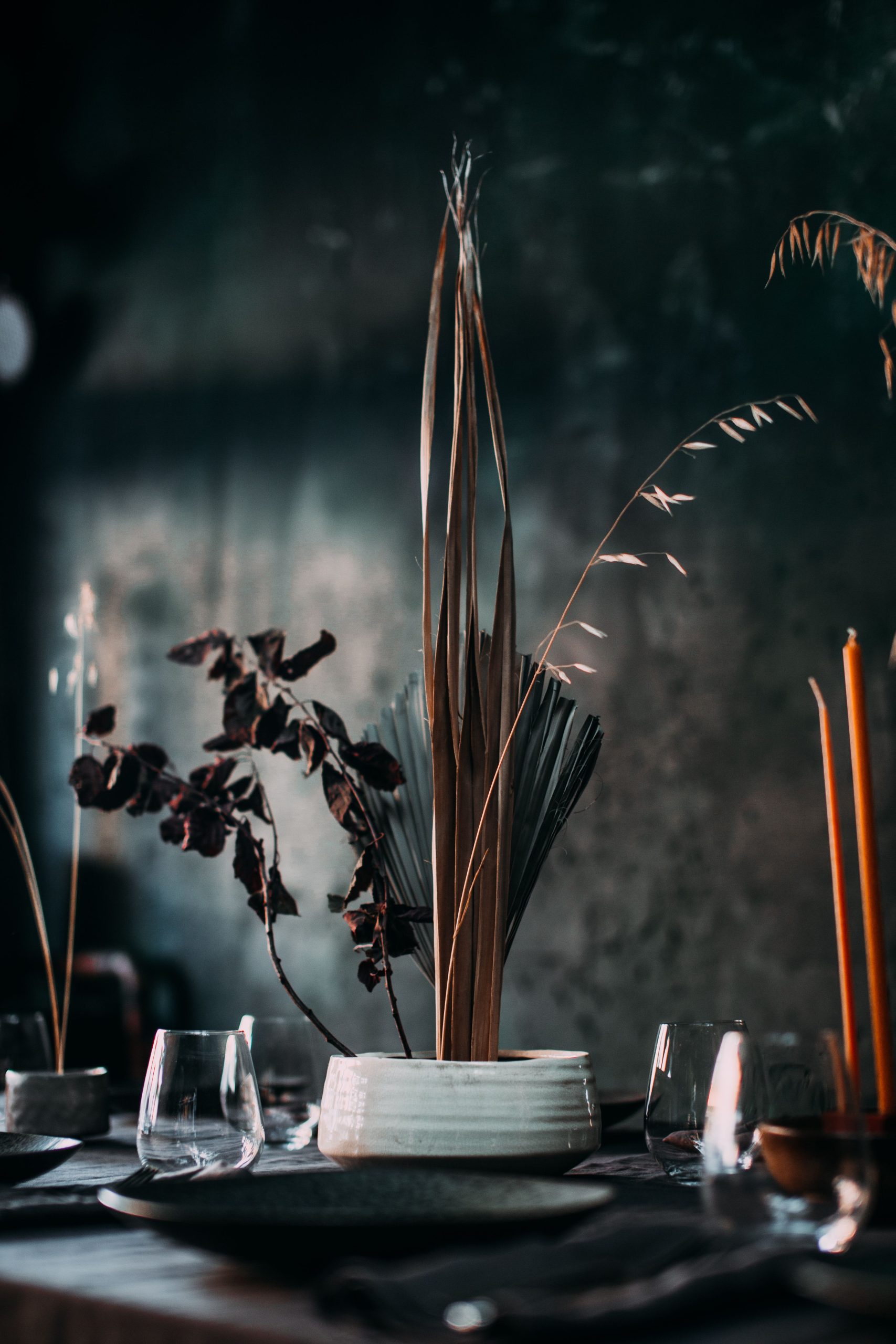 Composition of dry leaves and branches in white vase placed on table with dark tablecloth and utensil among candles and glasses with dark wall on background