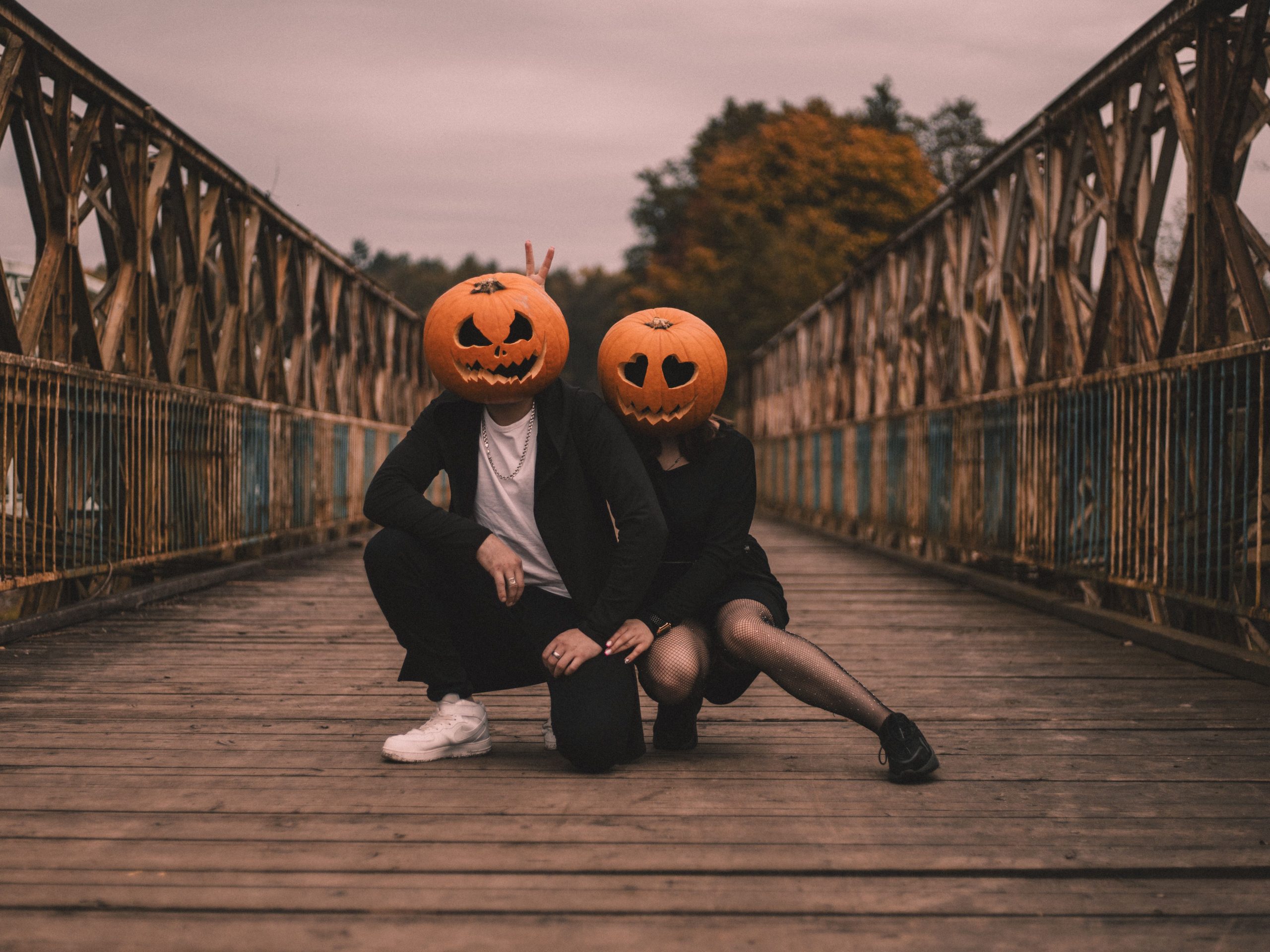 Couple in Halloween costumes with pumpkin heads on an old wooden bridge, adding a spooky charm to the scene.