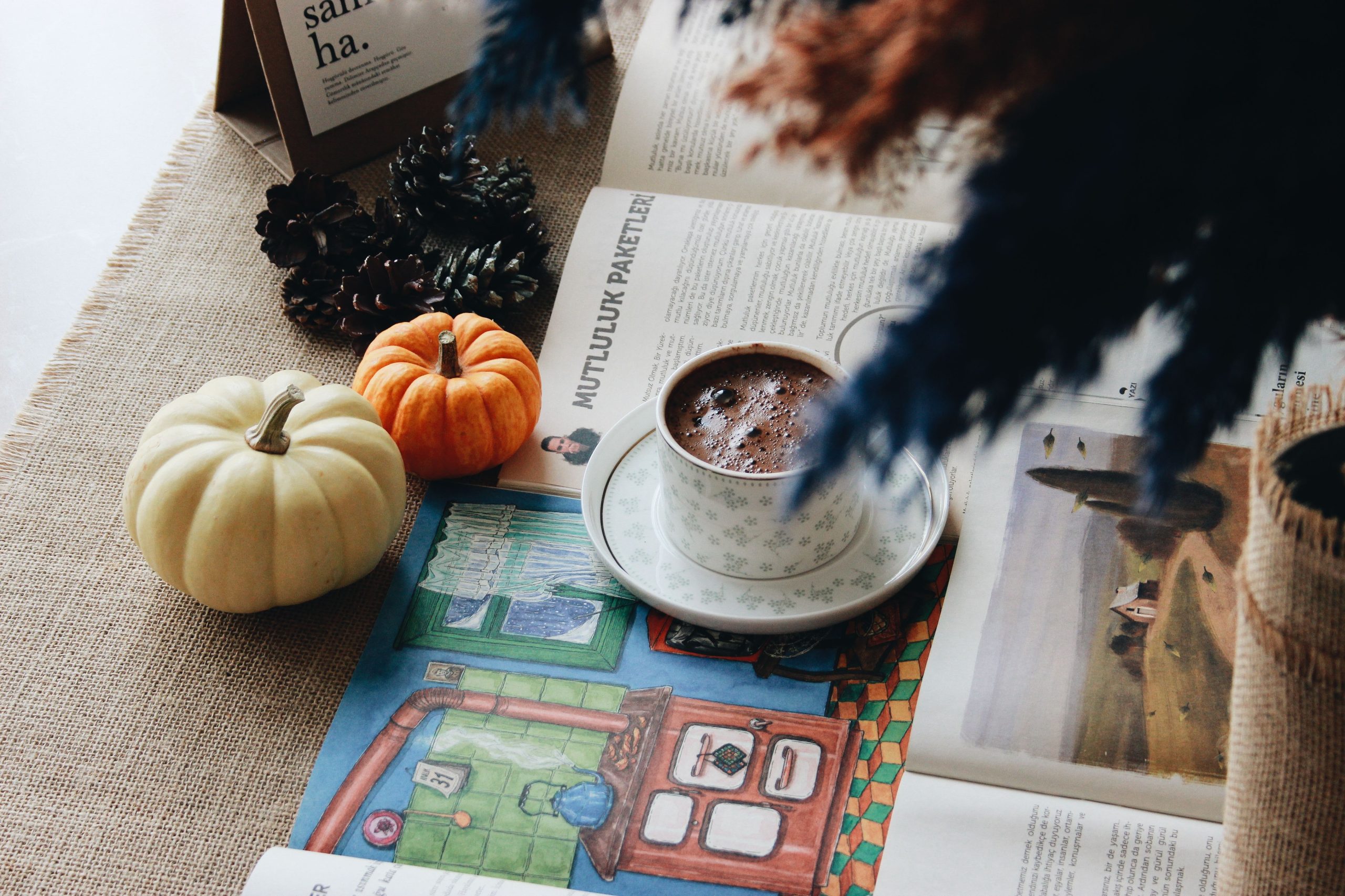 Cozy autumn setup with coffee, magazines, pumpkins, and pine cones on a rustic burlap backdrop.