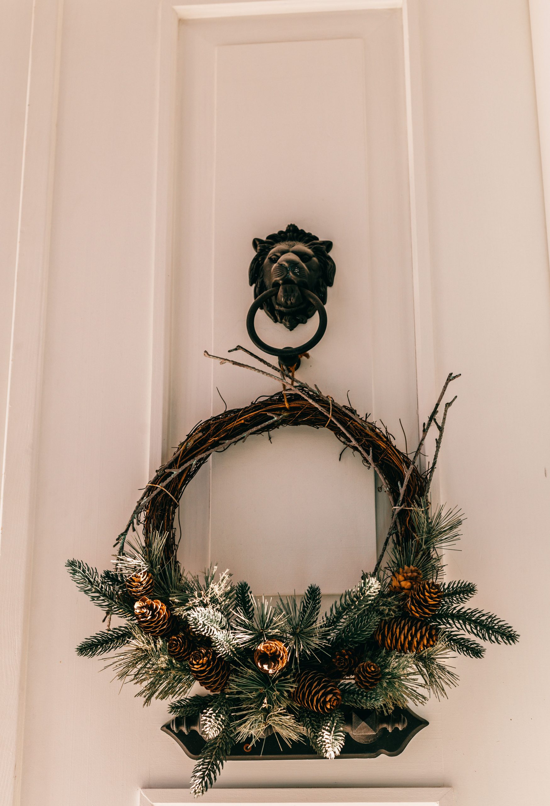 Decorative festive wreath with fir cones and twigs hanging on lion knocker on white front door