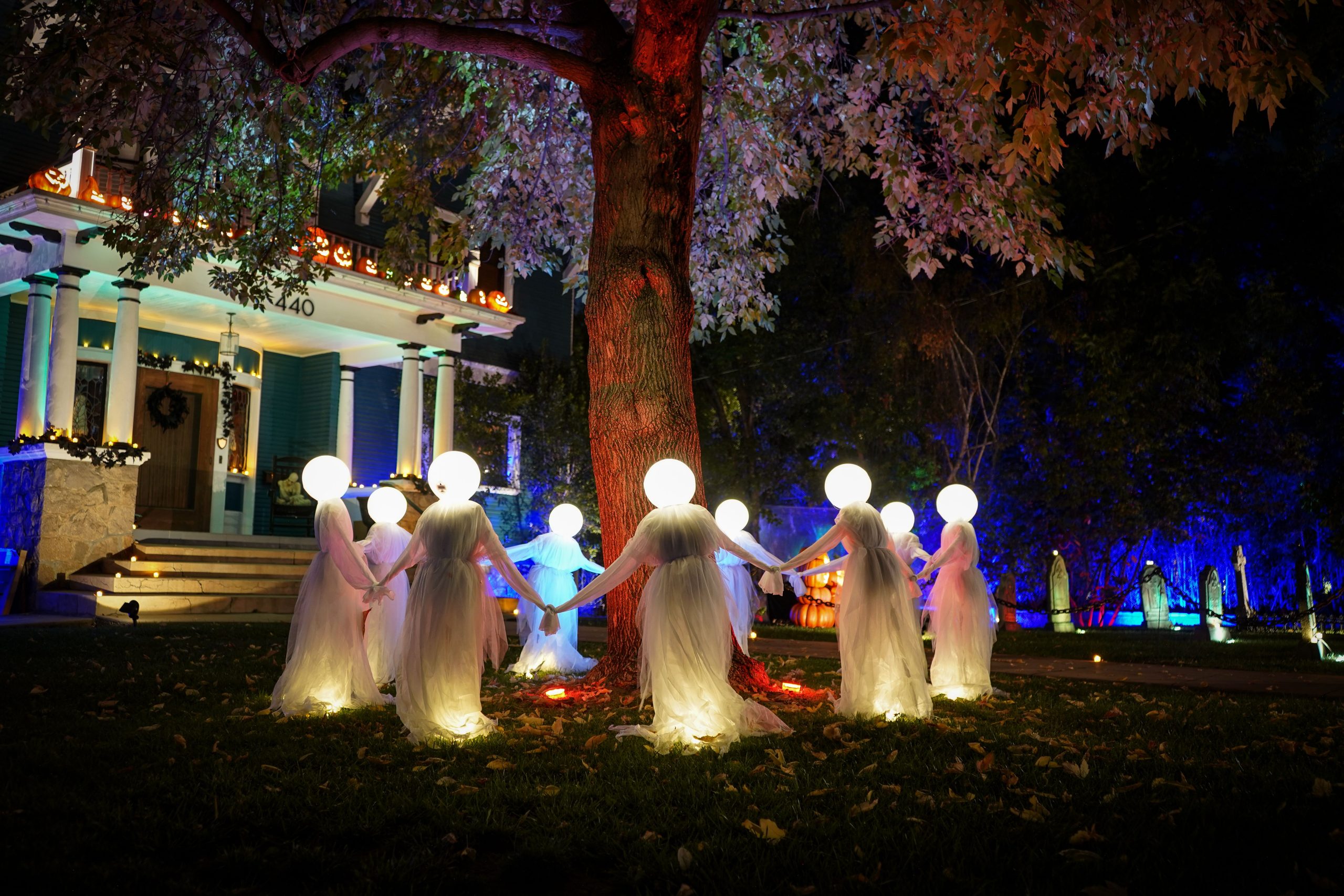Eerie Halloween display featuring illuminated ghost decorations under a tree at night in a suburban yard.