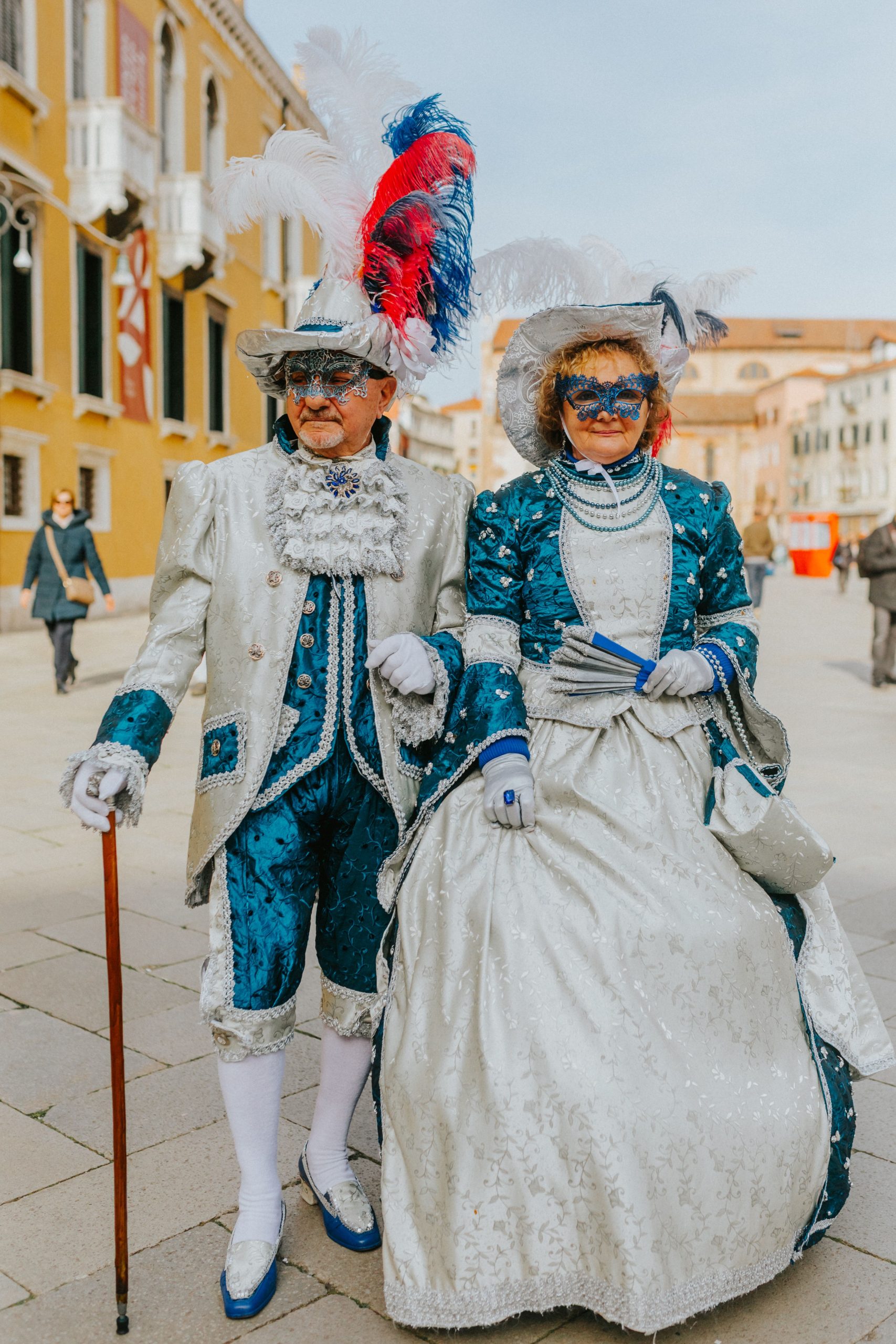 Elegant couple adorned in traditional Venetian carnival costumes strolls through picturesque Venice street.