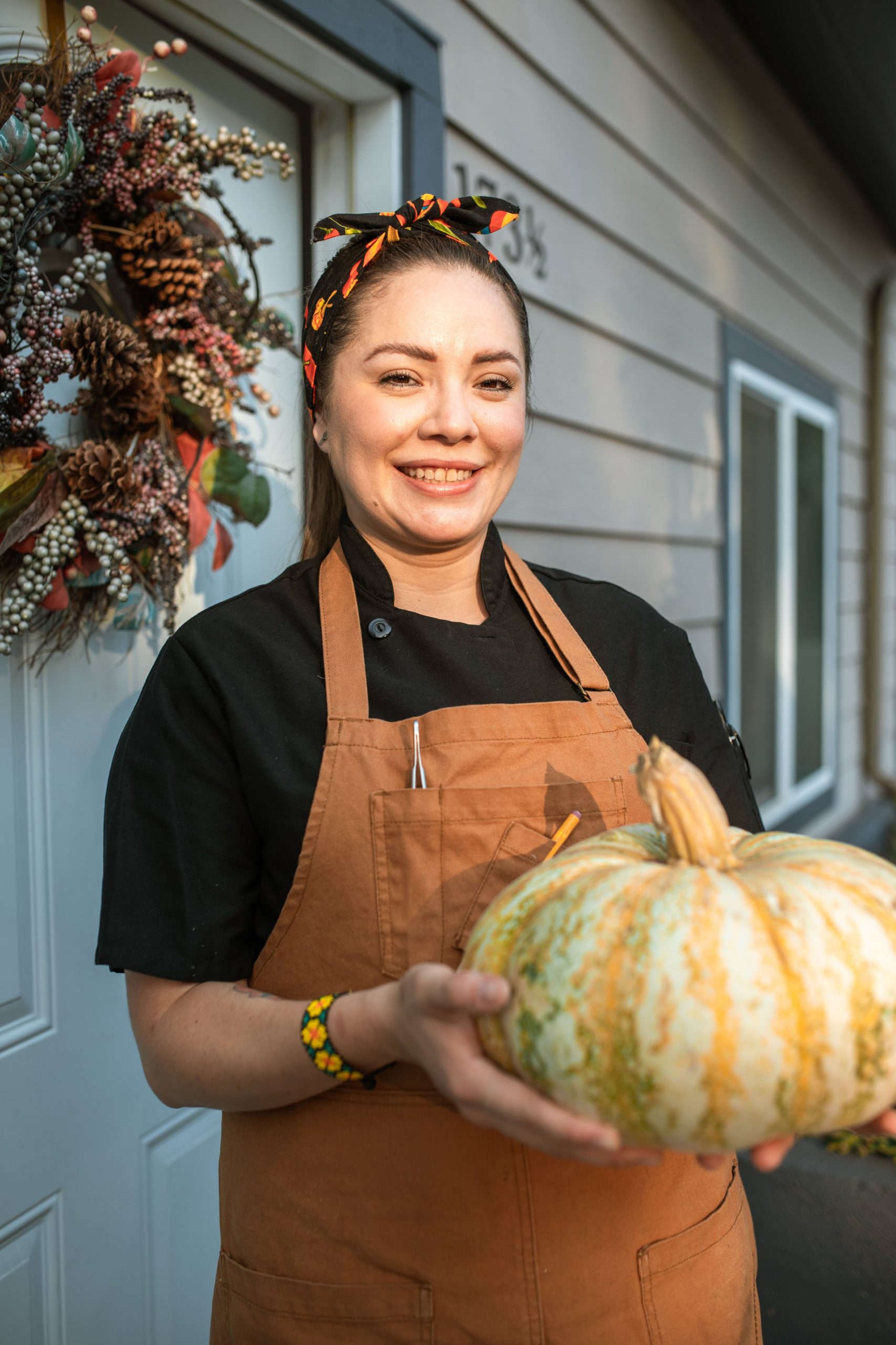 Happy woman in apron holding a pumpkin, standing by a wreath-decorated door, symbolizing autumn harvest.