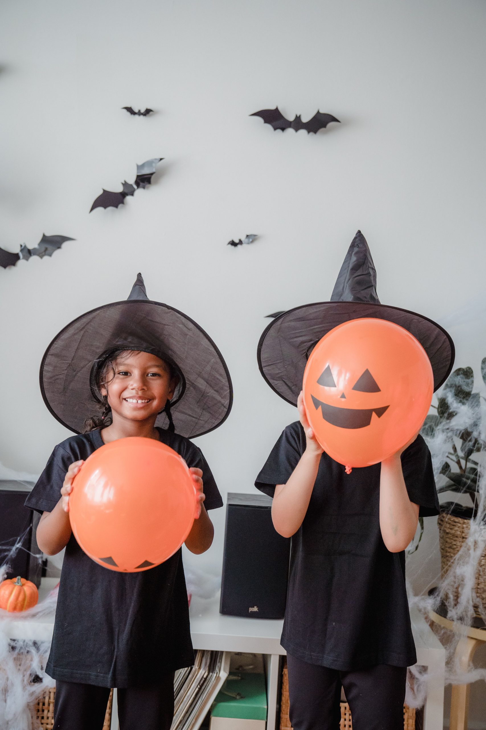 Kids dressed as witches holding orange balloons in a decorated Halloween setting.