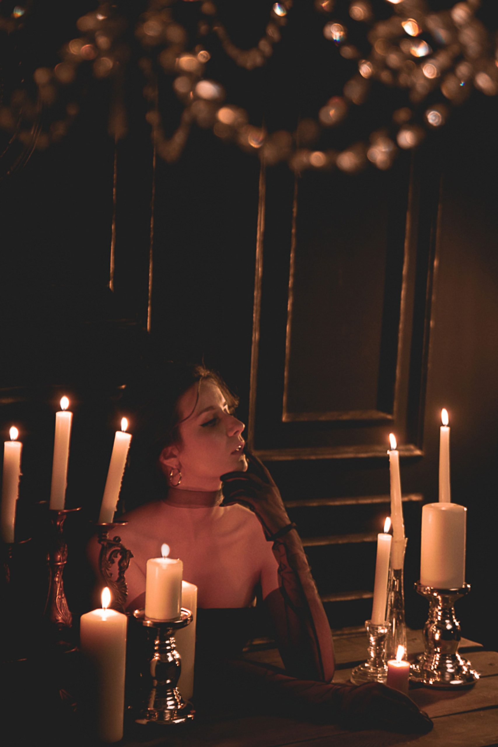 Lady in black clothes and gloves at wooden table with burning wax candles in dark house with ornamental walls and chandelier