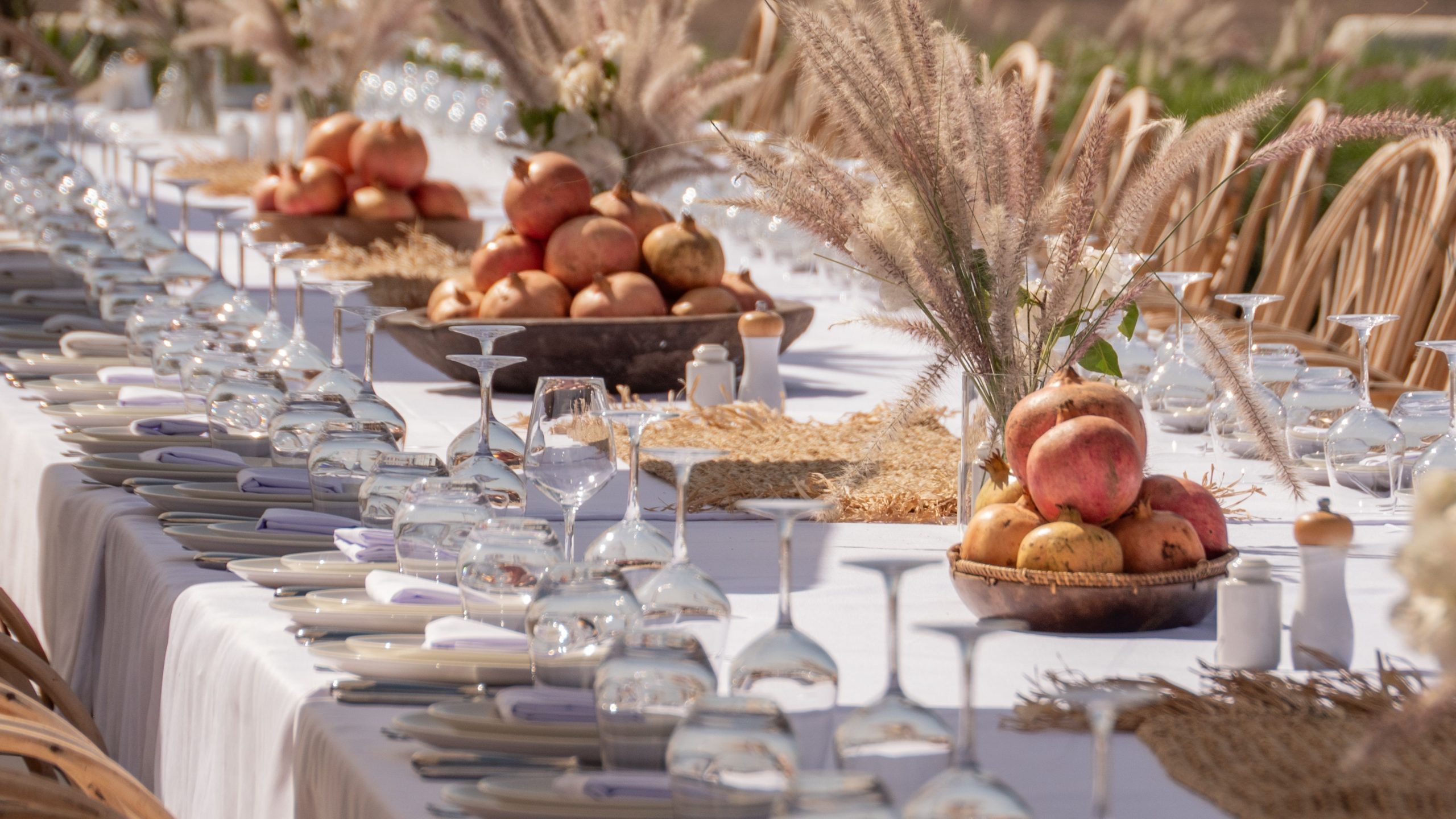 Long banquet table with elegant setup featuring pomegranates and glassware, perfect for an outdoor celebration.