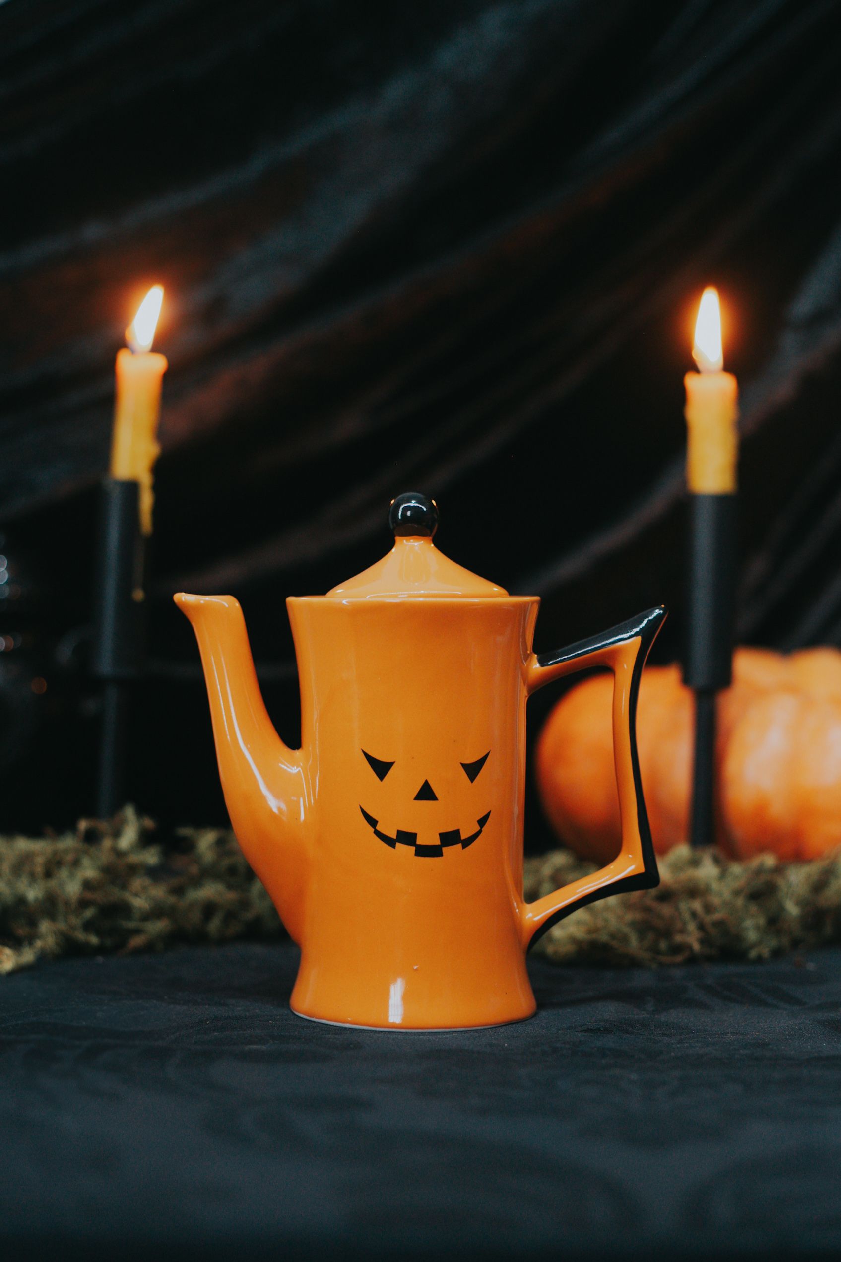 Orange teapot with a jack-o'-lantern face, surrounded by candles and pumpkins, perfect for Halloween celebrations.