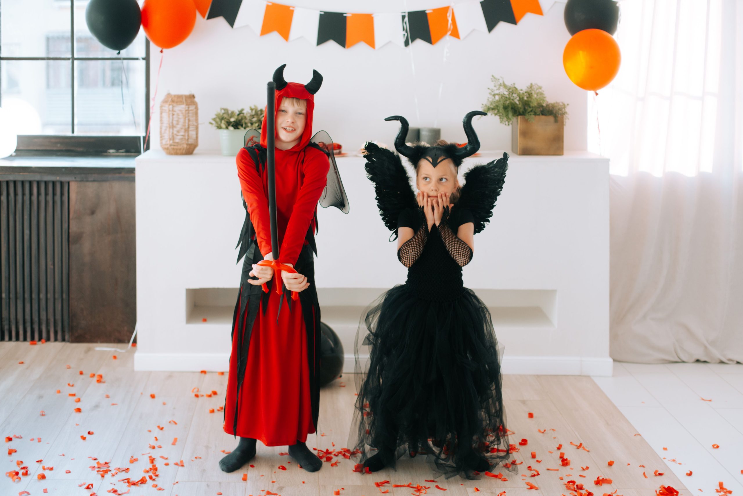 Two children in devil costumes celebrate Halloween indoors, surrounded by festive balloons.