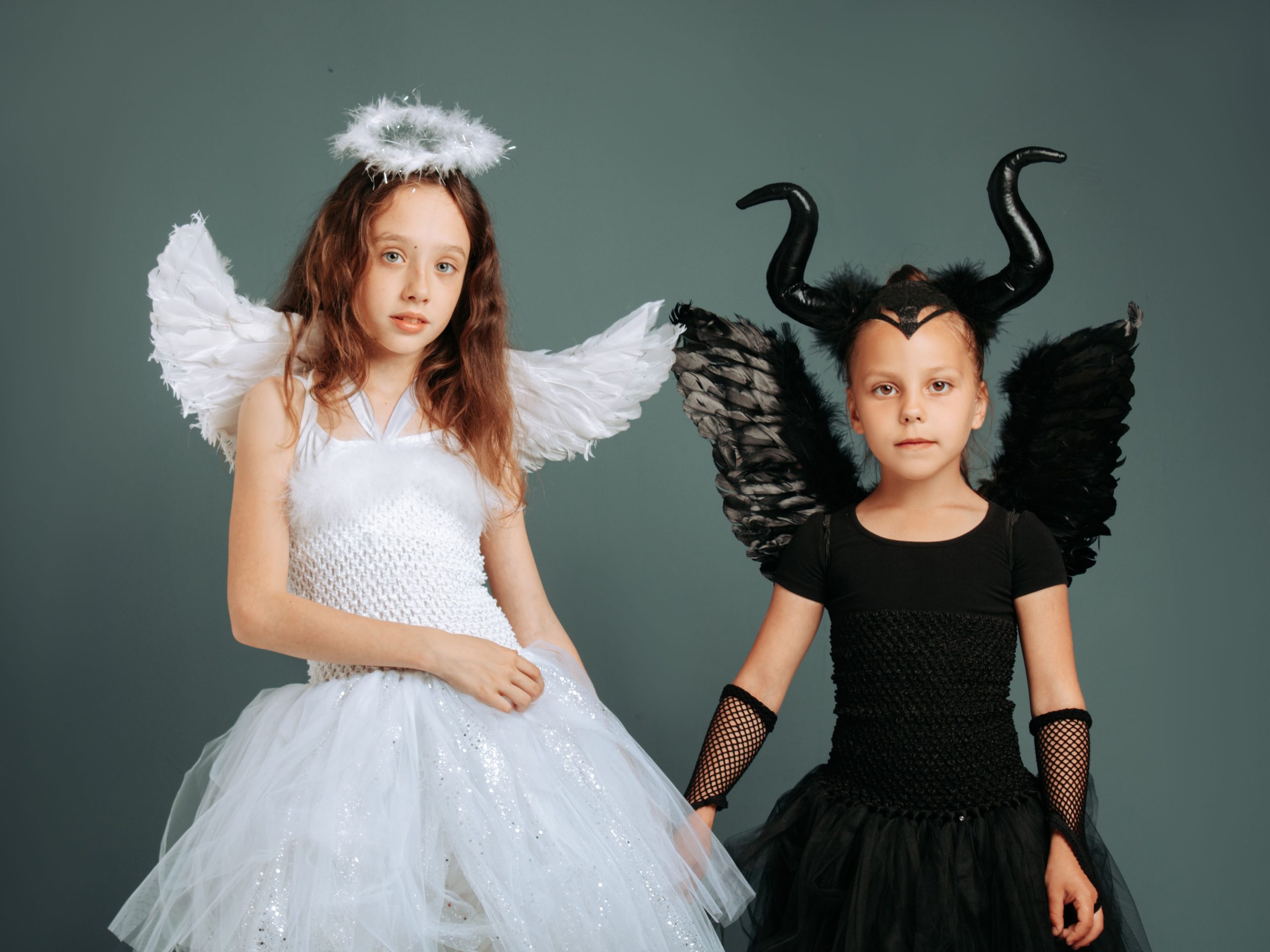 Two girls in angel and devil costumes with wings and horns against a gray background.