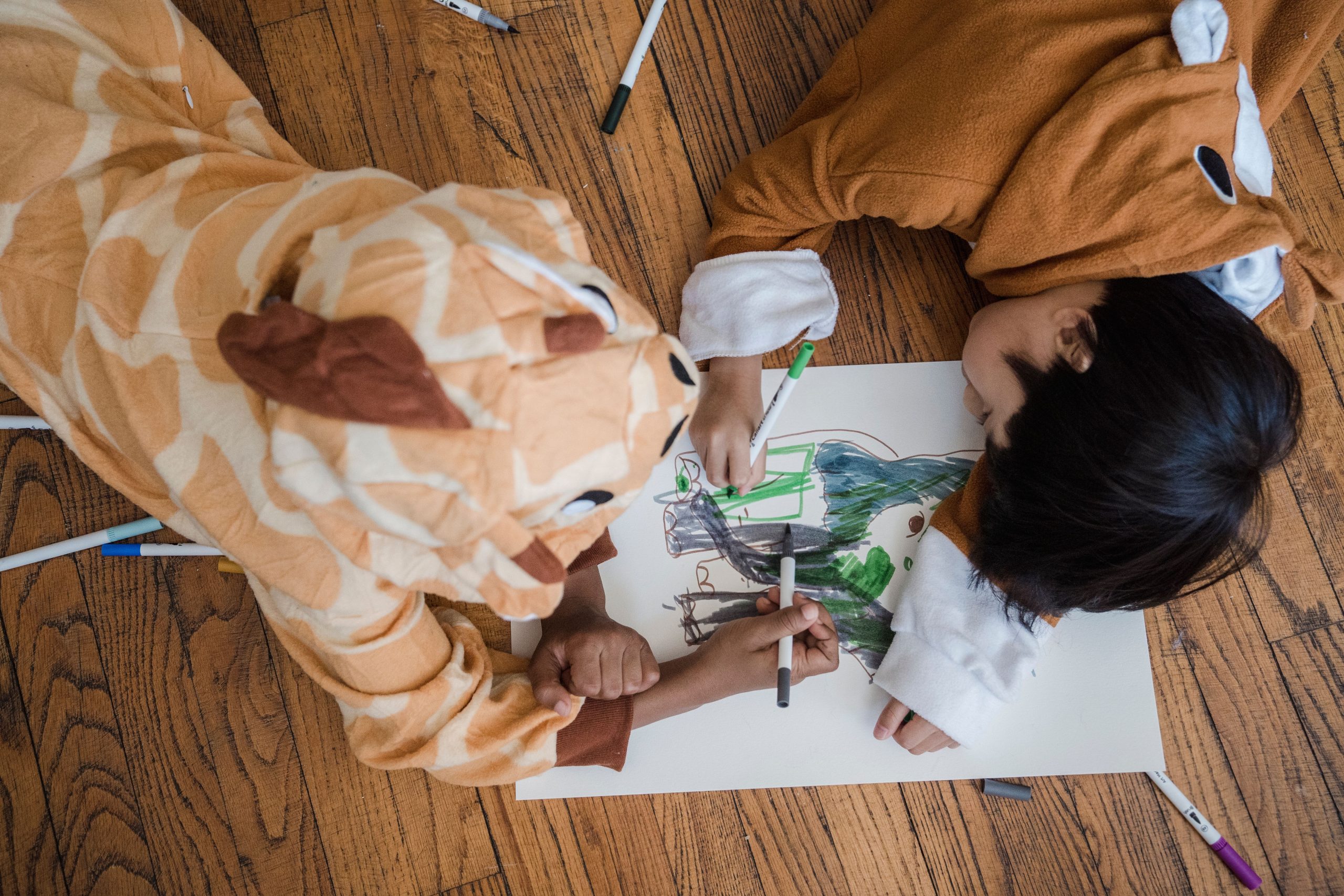 Two kids in animal costumes drawing with markers on a wooden floor.