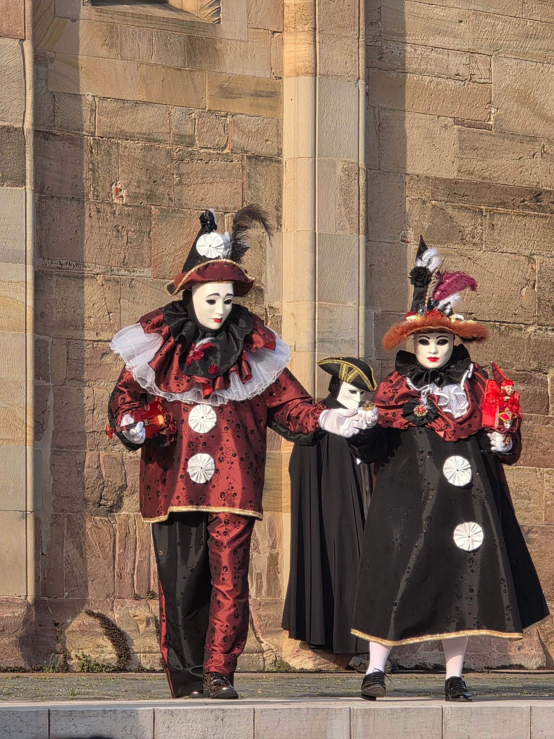 Two people in elaborate Venetian costumes and masks stand against a stone wall outdoors.