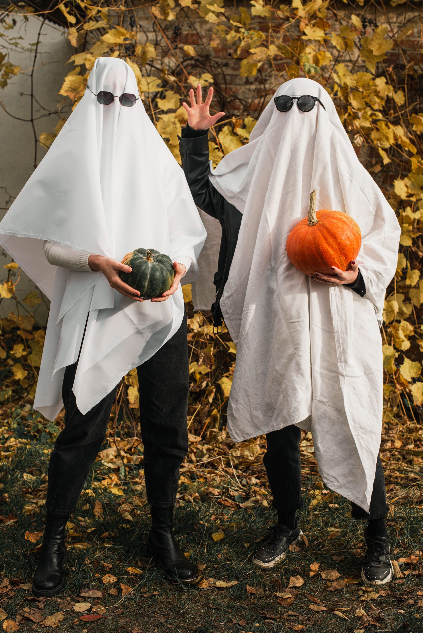 Two people in ghost costumes holding pumpkins amidst fall leaves outdoors.
