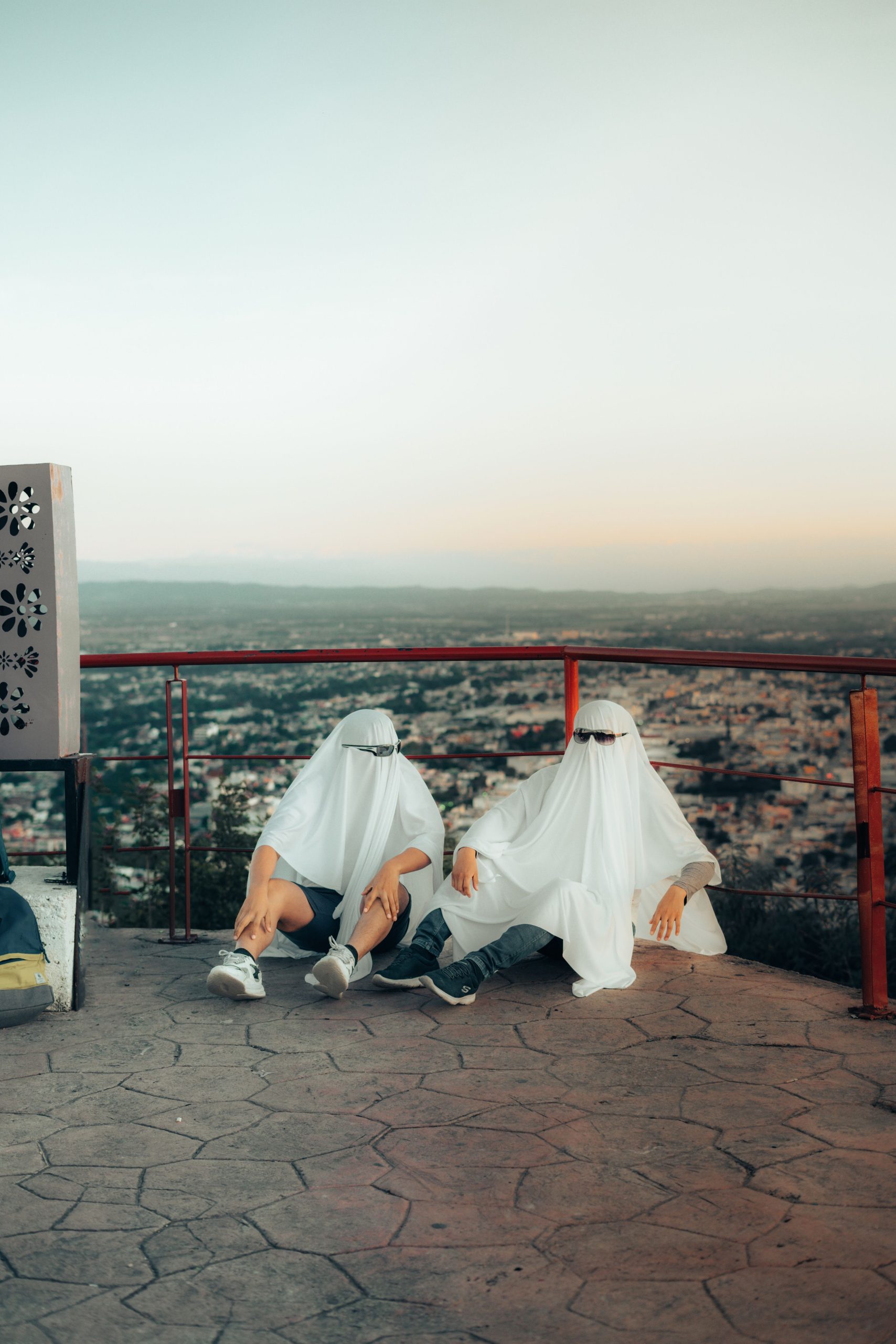 Two people in ghost costumes with sunglasses overlook a city from a scenic viewpoint.