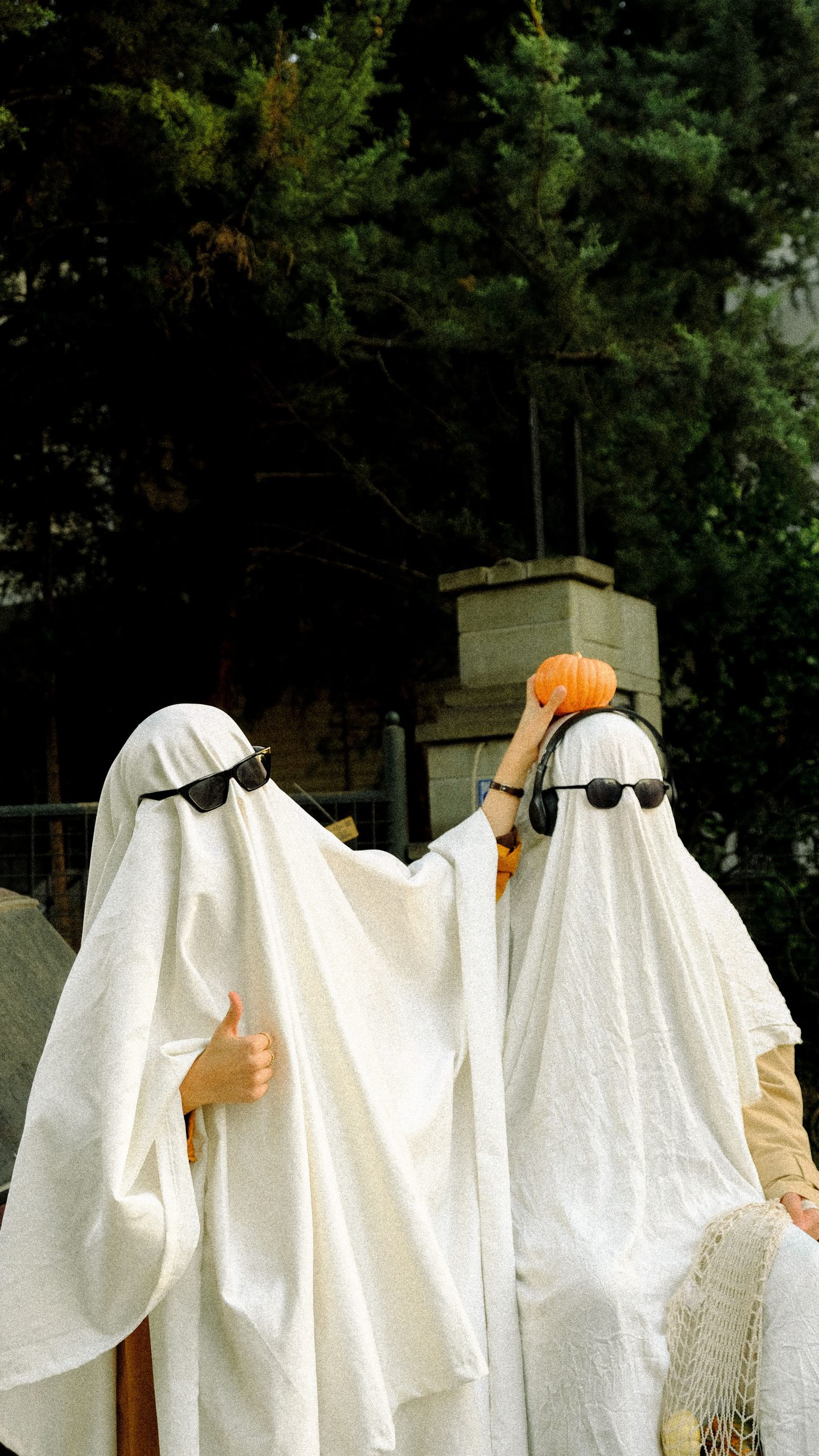 Two people in ghost costumes with sunglasses, holding a pumpkin outdoors.